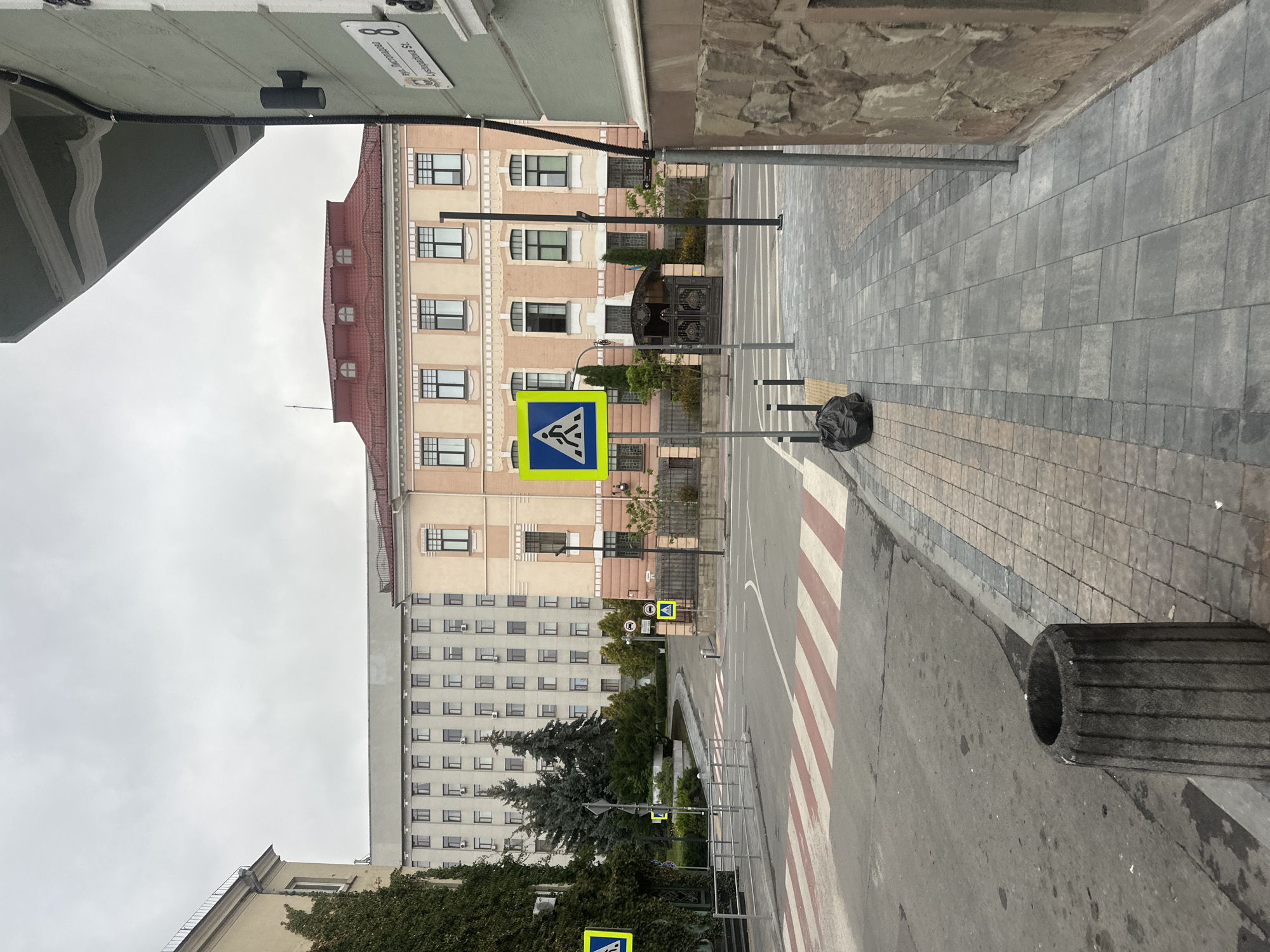 The image depicts a street scene in Ternopil, Ukraine, featuring a pedestrian crossing, buildings, and street signs. A prominent pedestrian crossing with red and white stripes is visible in the foreground, leading into a street with parked cars and various buildings. On the right side of the street, a light green building with a stone facade and a dark overhang is present. Above this building, a street sign with Cyrillic and Latin lettering is mounted, indicating "вул. Листопадова St. 8". In the background, a large, light-colored building with numerous windows stands behind a peach-colored building with a red roof. Trees and foliage are interspersed between the buildings and along the street. There are no people visible in the frame. The sky is overcast, suggesting cloudy weather.