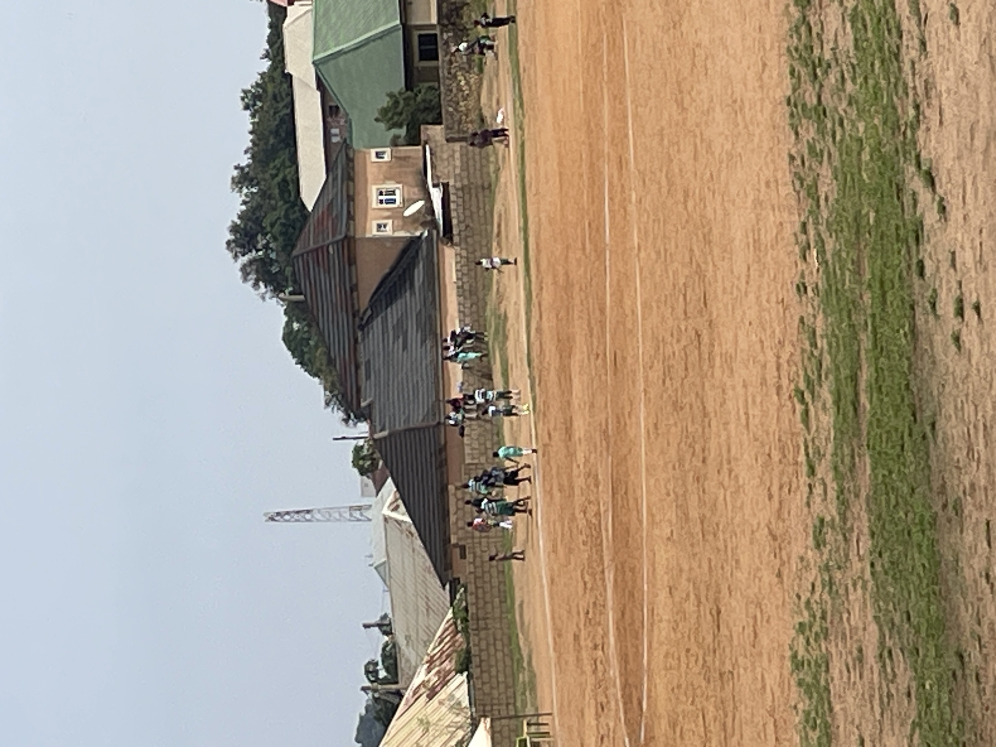 The image depicts an outdoor scene in Bukuru, Nigeria, likely a sports field or training ground. A dirt field occupies the foreground, with patches of sparse green grass visible. In the middle ground, a group of children and adults are gathered, some engaged in what appears to be a game or practice. Several individuals are wearing green and white attire, suggesting team participation. Behind the field, a brick wall separates it from buildings. These structures include houses with various roof types, some corrugated metal and others tiled. A communication tower is visible in the distance, along with lush trees and a light-colored sky. The overall impression is one of community activity within a residential urban or semi-urban setting.