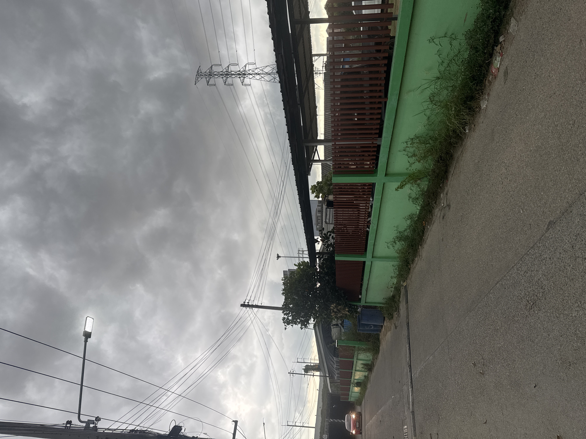 A cloudy day in Na Kluea, Thailand. The image, taken at ground level, depicts a street running alongside a long, green fence with brown wooden slats at the top. Overgrown vegetation grows along the base of the fence. Power lines and poles stretch across the sky. A tall, metal electricity pylon stands near the fence. A streetlight is on the left. A tree is near the fence. A car is visible in the distance. The street is made of asphalt. A blue trash can is by the fence.