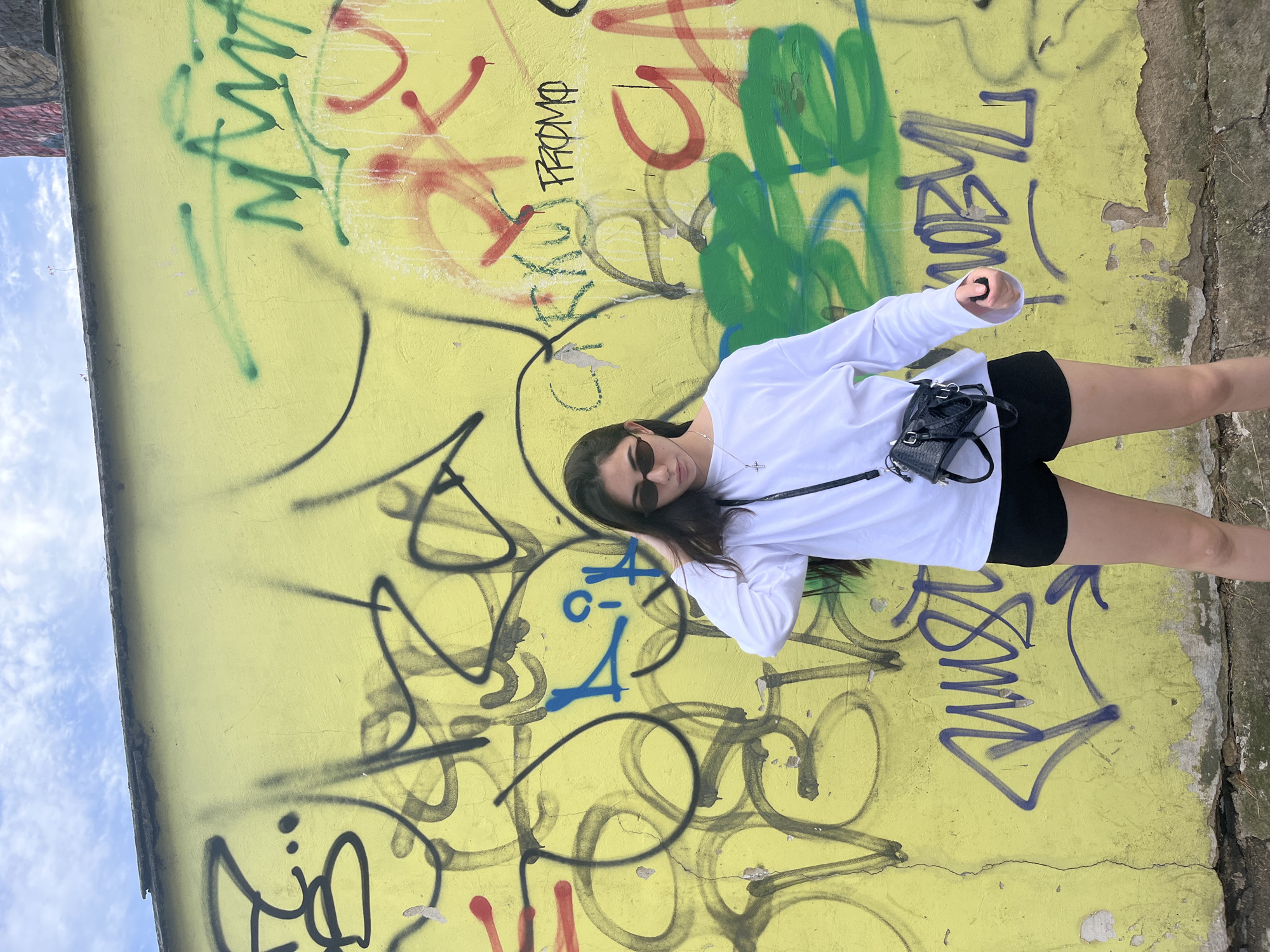 A young woman with long dark hair and sunglasses stands in front of a yellow wall covered in graffiti. She is wearing a white long-sleeved shirt, black shorts, and a small black purse. The graffiti on the wall is a mix of spray-painted tags and scribbles in various colors, including black, blue, green, red, and white. The wall appears to be an exterior wall of a building in Pilsen, Czechia. The sky above is partly cloudy.