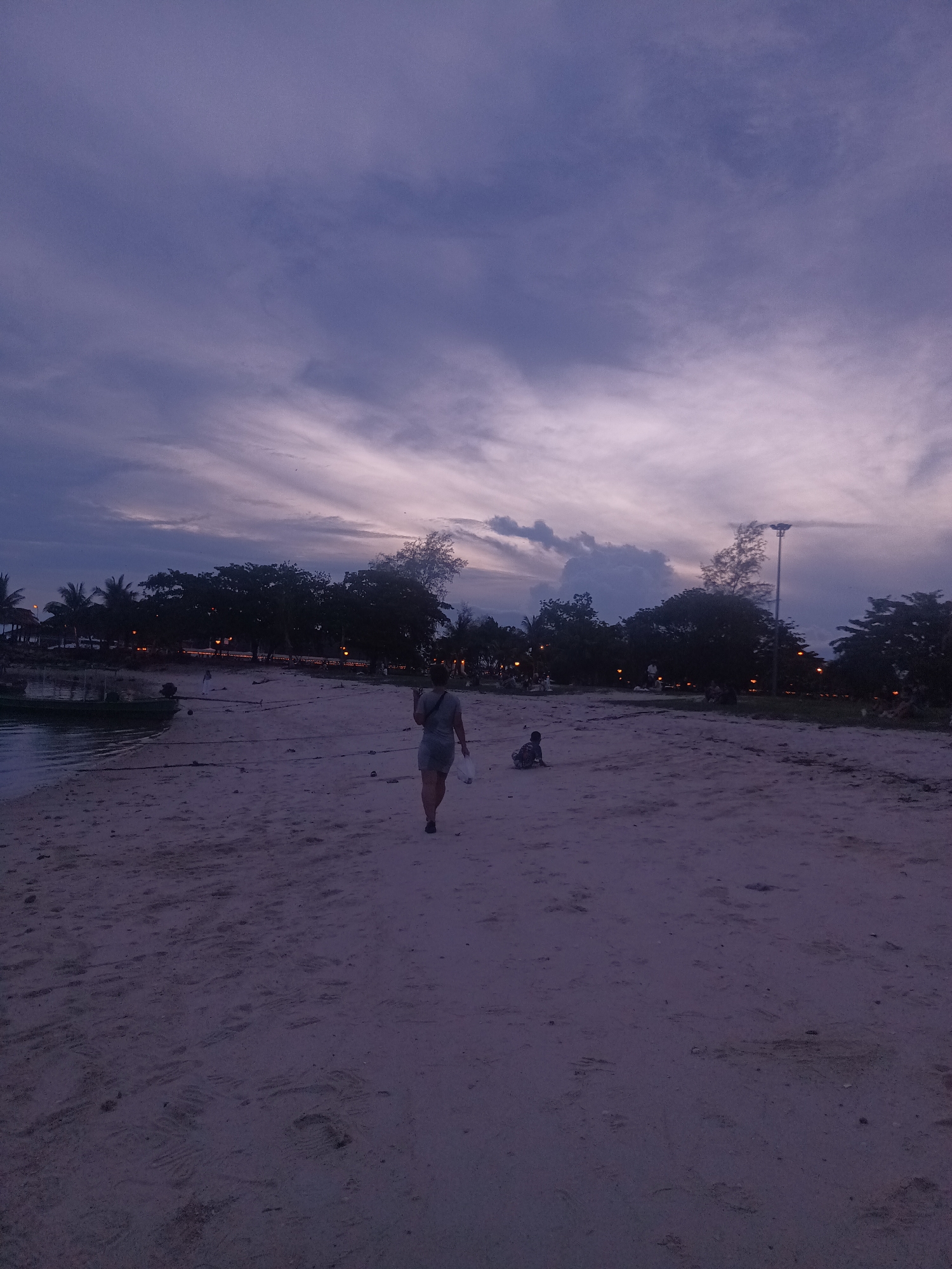 The image depicts a sandy beach scene in ตำบลเกาะพะงัน, Thailand, at dusk or twilight. The upper half of the frame is dominated by a cloudy sky exhibiting a gradient from deep indigo at the zenith to pale purple and pink hues near the horizon, with visible cloud streaks. A dark, irregular line of dense foliage, consistent with tropical trees, separates the sky from the land. Embedded within and behind this foliage, numerous small, warm-toned artificial lights are visible, suggesting a developed area beyond the immediate beach. On the far right, a tall, dark streetlight pole rises above the tree line.
The foreground and midground consist of light-colored sand, appearing muted gray-purple under the low light, with discernible footprints. To the far left, a calm body of water reflects the dim sky. A small, dark boat, equipped with an outboard motor, is partially afloat and partially resting on the sand near the water's edge, secured by ropes extending onto the beach.
Two individuals are present on the beach. One adult figure, with their back to the camera, walks towards the left mid-ground, wearing light-colored attire and carrying a white bag in their right hand. Further to the right in the mid-ground, a smaller individual, likely a child, is seated directly on the sand, facing away from the camera and towards the line of trees. The overall scene conveys a tranquil, low-light environment with minimal human activity.