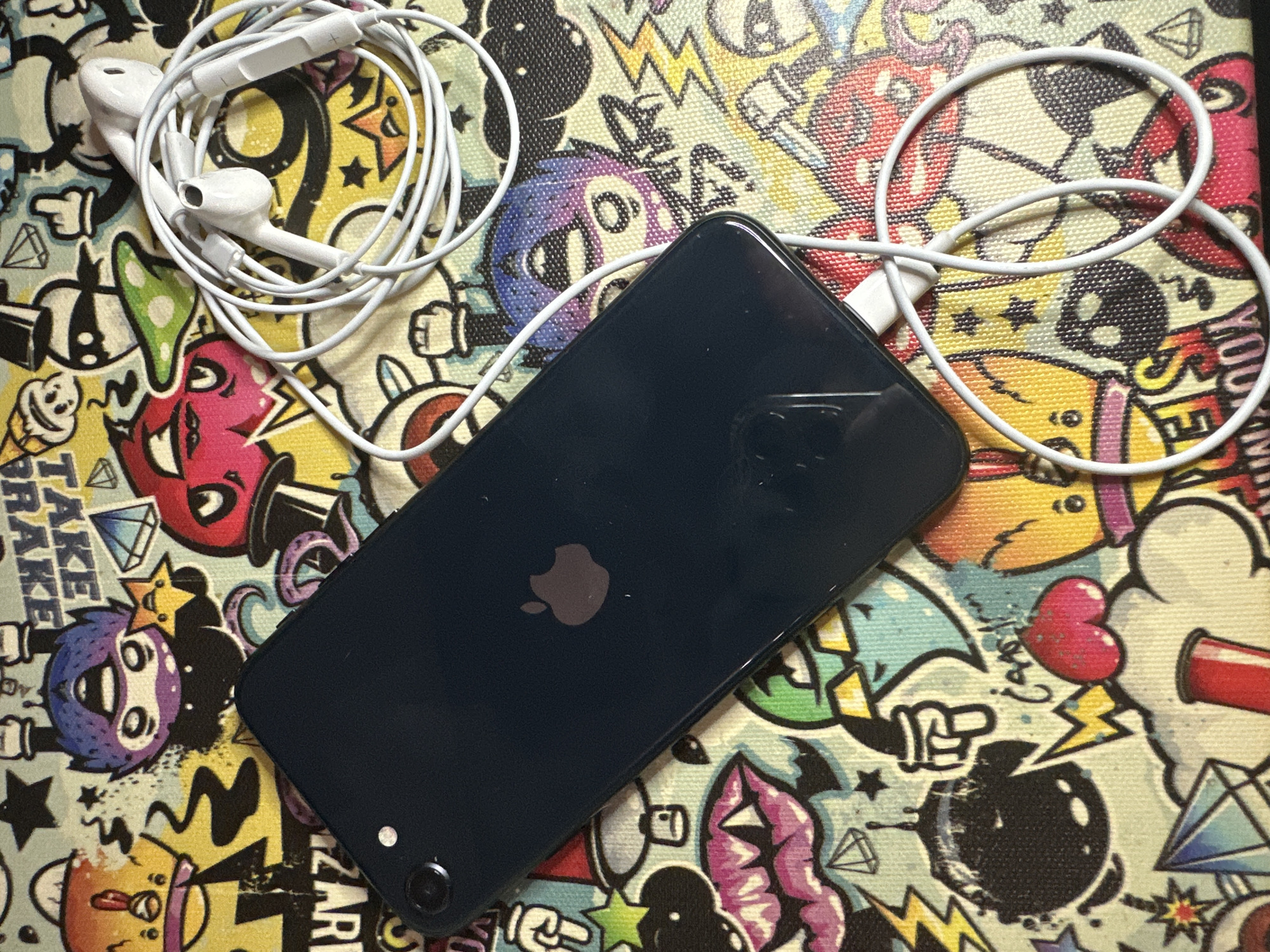 The image shows a dark blue iPhone lying on a colorful, cartoon-style patterned surface. Next to the iPhone are white wired earbuds, coiled up. The iPhone's screen is not visible, but the back of the phone, including the Apple logo, is. The background is busy with various illustrated characters and designs, including stars, lightning bolts, and cartoon faces. The scene is brightly lit and appears to be a flat lay arrangement of personal electronic items. There is no indication of the location being Moscow, Russia.