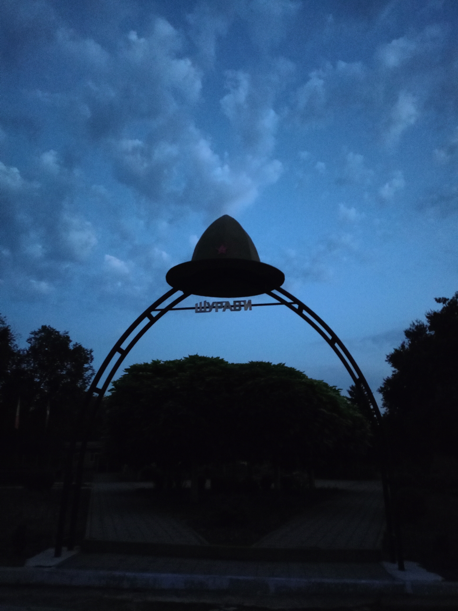 An outdoor, low-light, vertical photograph captures a silhouette of an arched entranceway topped with a large, conical hat-like structure featuring a red star insignia. The hat is suspended above the archway, which is adorned with Cyrillic lettering. The scene is set against a dark blue, cloudy sky. Silhouetted trees and bushes are visible behind the archway, framing a paved pathway that recedes into darkness. The overall impression is one of dusk or twilight, with limited visibility and a somber atmosphere. The location is identified as Baurci, Moldova.