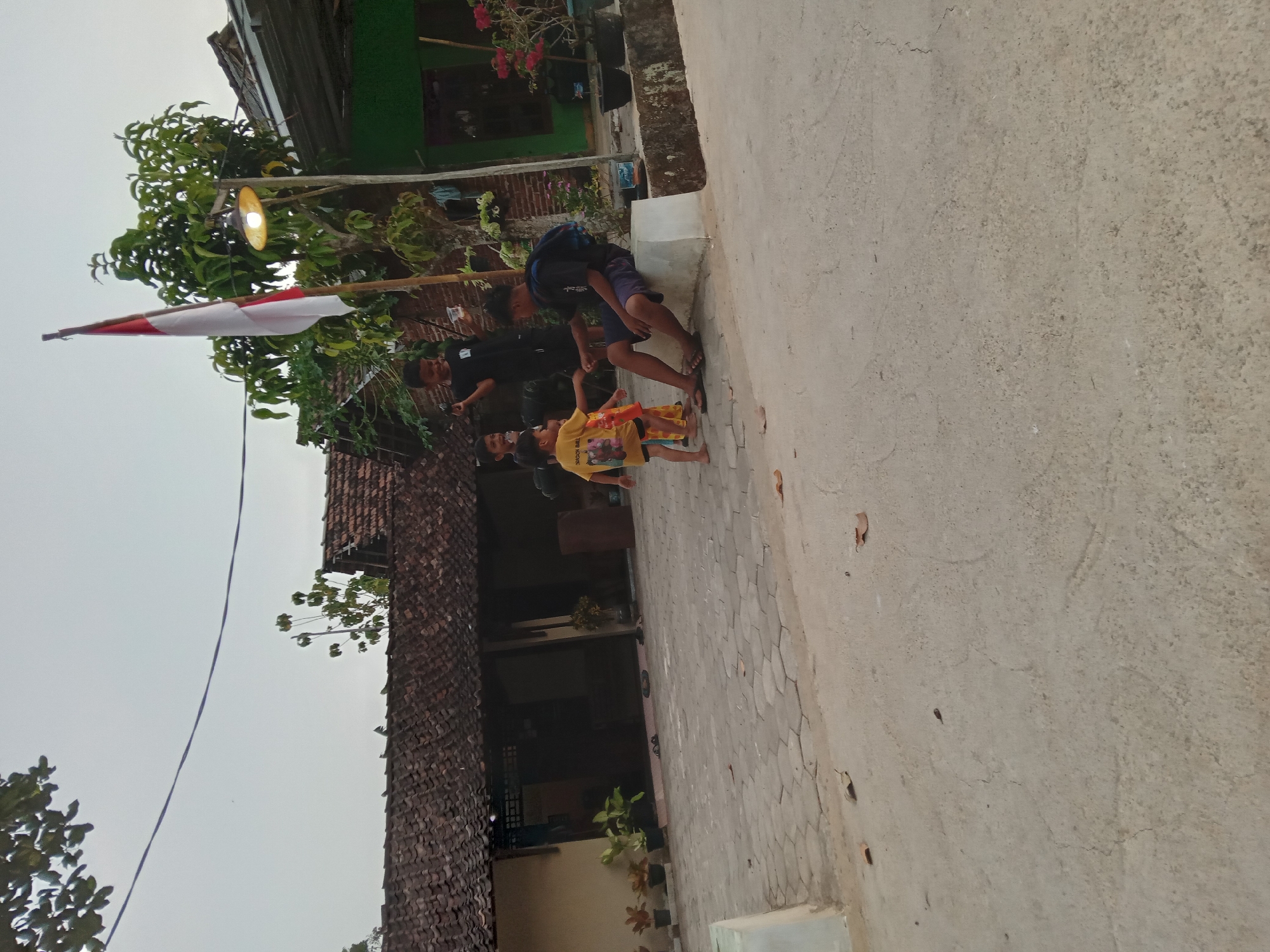 In Kedunggudel, Indonesia, the image captures a scene in front of traditional Indonesian homes. The foreground shows a concrete pathway, weathered with cracks and scattered with dry leaves. To the left, a raised white structure is visible. Behind this, a paved courtyard made of hexagonal tiles leads to the houses, characterized by their dark, tiled roofs. Several potted plants with green foliage are placed along the side of the houses.
In the mid-ground, five young boys are interacting. One boy, wearing a yellow t-shirt with a "Dragon Ball" graphic, stands facing a boy sitting on a low white concrete ledge. The sitting boy, dressed in a dark t-shirt and shorts, is looking towards the standing boy and appears to be holding his hand. Behind them, three other boys are gathered. One boy in a black t-shirt is looking towards the left, holding a clear plastic cup. Another boy, also in black, is looking forward. A fourth boy, partially obscured, is standing near the other children.
A flagpole with the Indonesian flag, red on top and white on the bottom, stands prominently to the right of the group of children, positioned between a large tree with lush green leaves and a lamp post with a lit yellow bulb. The flag appears to be slightly tattered. Beyond the houses, a green building with a corrugated roof is visible, and in the background, potted plants with pink flowers add a touch of color. The sky is overcast and light.
