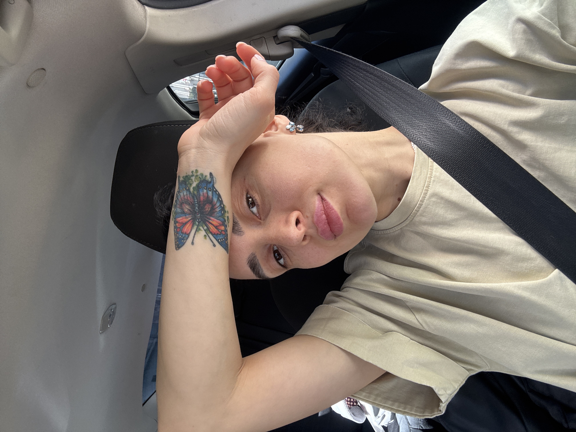 A person is seated in a car, wearing a seatbelt and a beige t-shirt. They have a butterfly tattoo on their left forearm. Their face is visible; they have light skin and are looking at the camera. The car interior is visible, with the dashboard and headrest in view. The location is assumed to be Tbilisi, Georgia. The scene appears to be a selfie taken inside a vehicle.