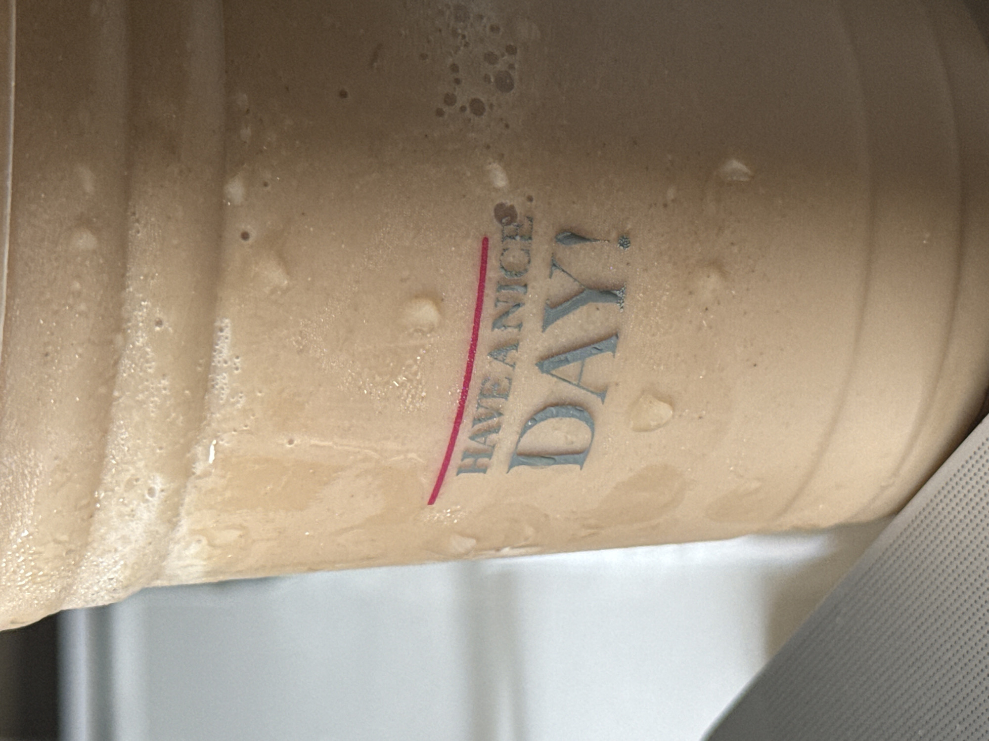 A close-up, slightly rotated image shows a light brown, likely chilled or cold, beverage in a bottle, possibly plastic, bearing the phrase "HAVE A NICE DAY!" printed on its surface in red and gray. Condensation is visible on the bottle, suggesting it contains a cold liquid. The lower portion of the image shows blurry parts of a light-colored, possibly white or gray, surface, perhaps a table or counter. The scene does not contain sufficient information to determine a specific location in Mexico City, Mexico, or details about people, actions, or interactions.