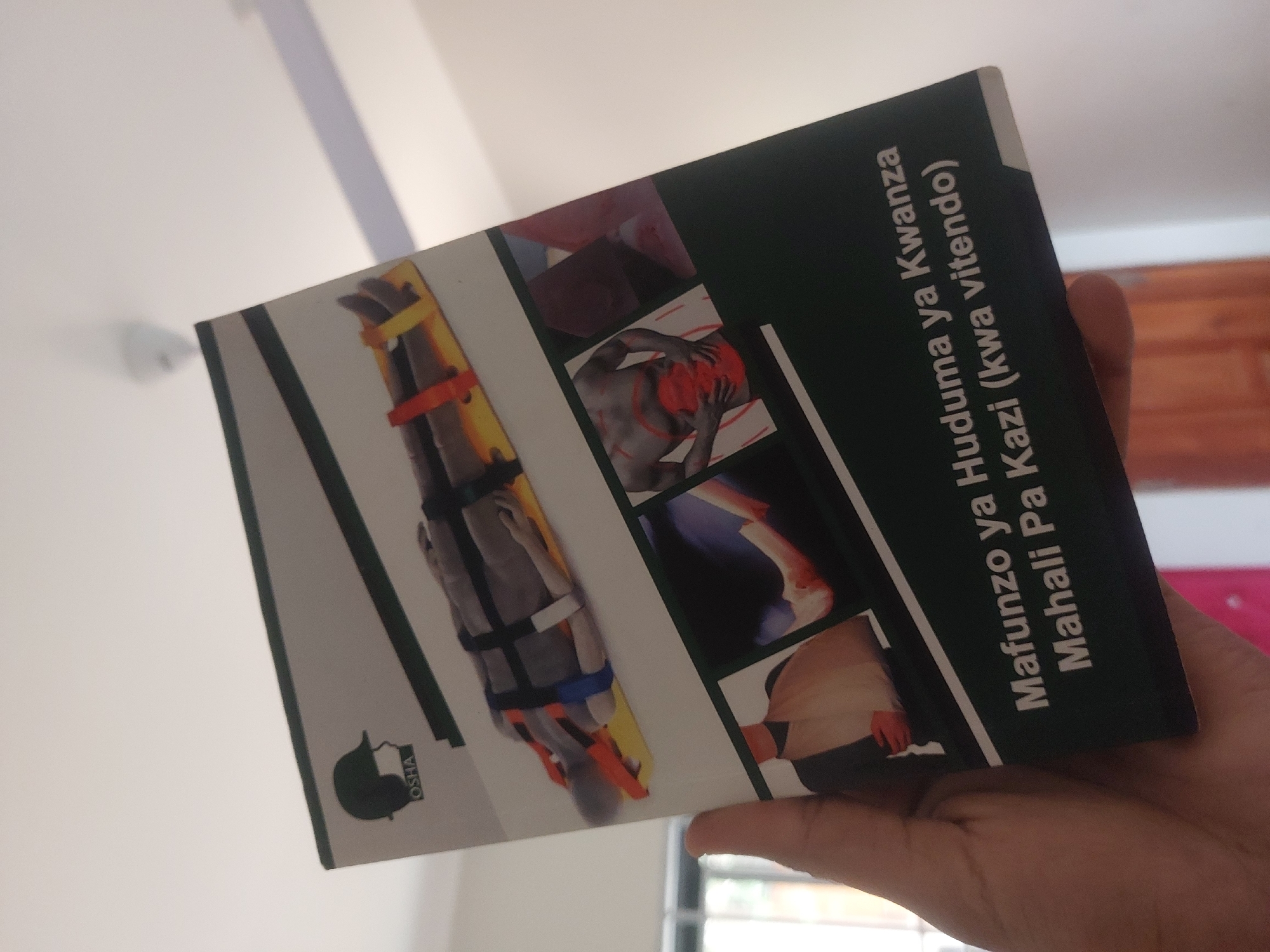 A person holds a book in their hand. The book is titled "Mafunzo ya Huduma ya Kwanza Mahali Pa Kazi (kwa vitendo)" which translates to "First Aid Training in the Workplace (in practice)." The cover features images related to first aid. An OSHA logo is visible on the book. The background shows a room interior with light walls. It is not possible to discern whether the photo was taken in Dar es-Salaam or anywhere in Tanzania, as there is no identifiable visual information indicating the location.