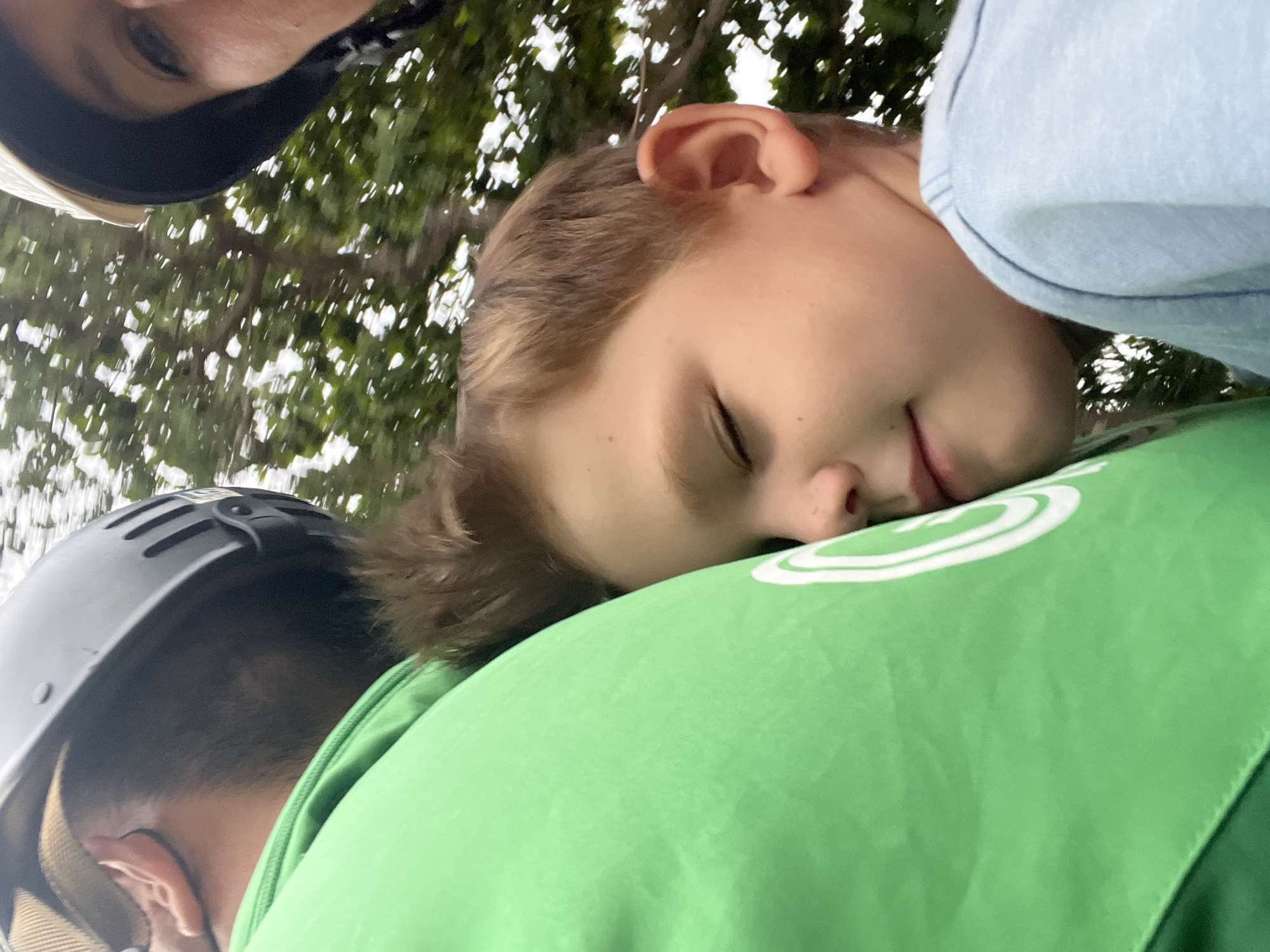 The image shows a close-up of a young boy sleeping, nestled against a person wearing a bright green shirt. The boy has closed eyes and his head is resting on the shoulder of the person in green. Another person, wearing a light-colored helmet, is visible in the upper right corner of the frame. The background is blurred, with trees and foliage suggesting an outdoor setting. The overall impression is one of a peaceful moment, possibly during travel. The specific location in Hoi An, Vietnam, cannot be determined from the visual information provided.