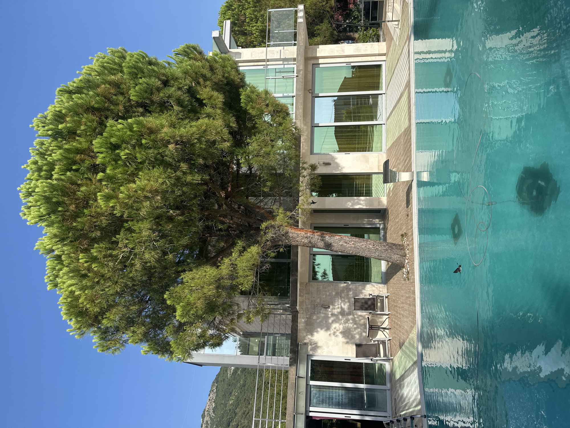 A tranquil scene unfolds in Brca, Montenegro, where a modern, multi-story house, constructed with stone and glass, stands majestically beside a sparkling turquoise swimming pool. The house features large glass windows that reflect the lush green surroundings and the clear blue sky. A mature pine tree, its branches reaching out like welcoming arms, is a prominent feature in the foreground, casting dappled shadows on the tiled patio. Two lounge chairs and a small table are placed on the patio, inviting relaxation. The swimming pool, with its smooth surface, mirrors the house and the sky, with a pool cleaning hose visible submerged in the water. In the distance, rolling green hills rise against the backdrop of a cloudless sky. The overall atmosphere is one of serene luxury and peaceful retreat.