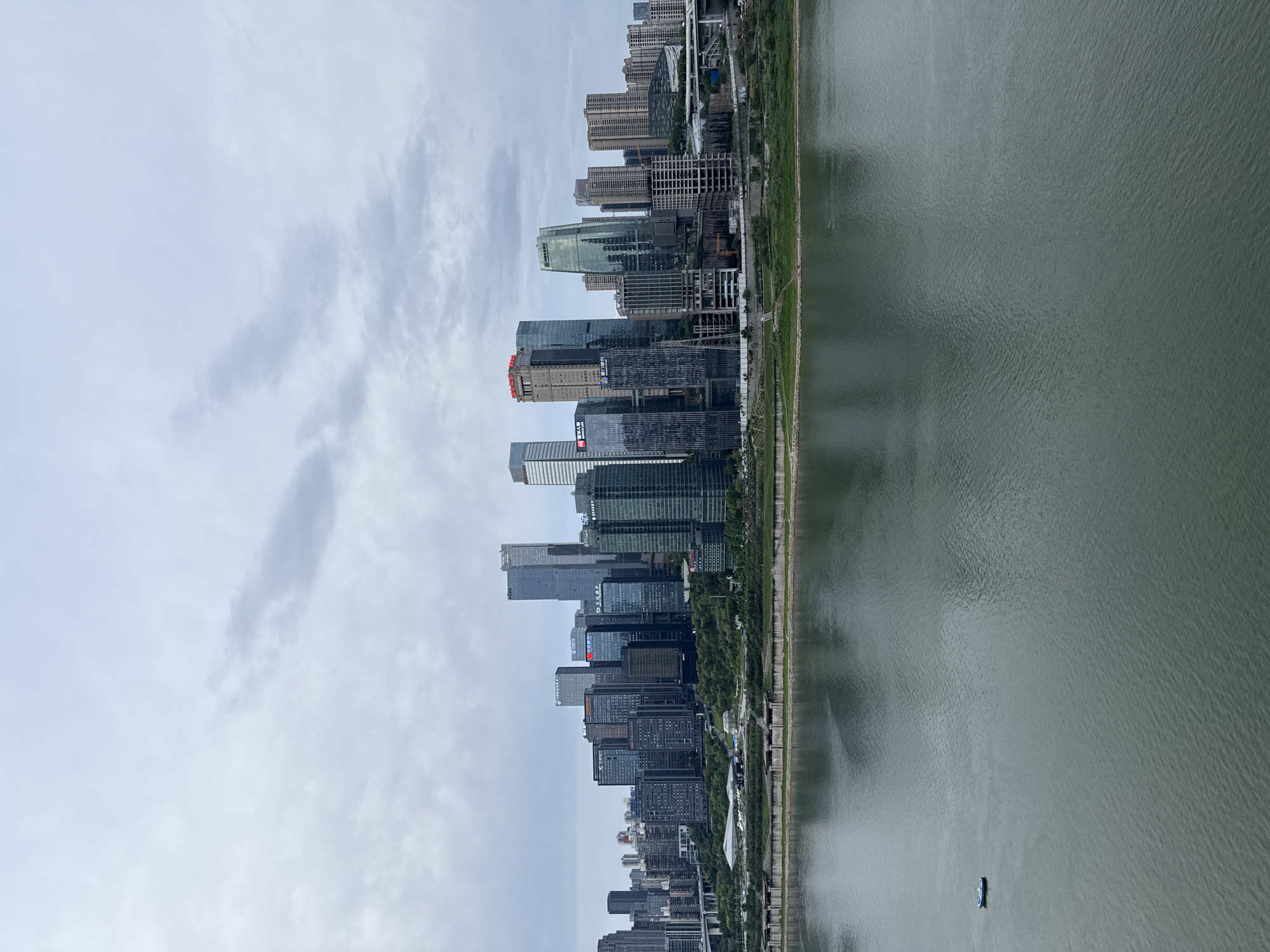 The image captures a panoramic view of a modern city skyline situated alongside a wide river, likely taken from an elevated position looking across the water towards the urban landscape.
The upper portion of the frame features a bright, overcast sky with diffused clouds, indicating a cloudy daytime setting without direct sunlight.
The central and dominant section of the image showcases a densely packed cluster of high-rise buildings and skyscrapers that line the riverbank. These structures exhibit a variety of modern architectural styles, many with reflective glass facades. Some buildings appear to be under construction, with visible frameworks and cranes suggesting ongoing development. A strip of green vegetation, likely trees or parkland, separates the buildings from the river's edge. Several buildings display prominent corporate logos and Chinese characters. Visible text includes red characters on one building that read "中国人民" (Zhōngguó Rénmín), and a red square logo on another, likely indicating a financial institution or large corporation.
The lower part of the image is filled by the wide expanse of the river, characterized by its calm, greenish-grey surface with subtle ripples. A small, dark-colored boat is visible on the water, suggesting either recreational use or commercial activity.
The scene depicts a bustling urban environment typical of a major Chinese metropolis, consistent with the specified location of Yuzhong District, China.