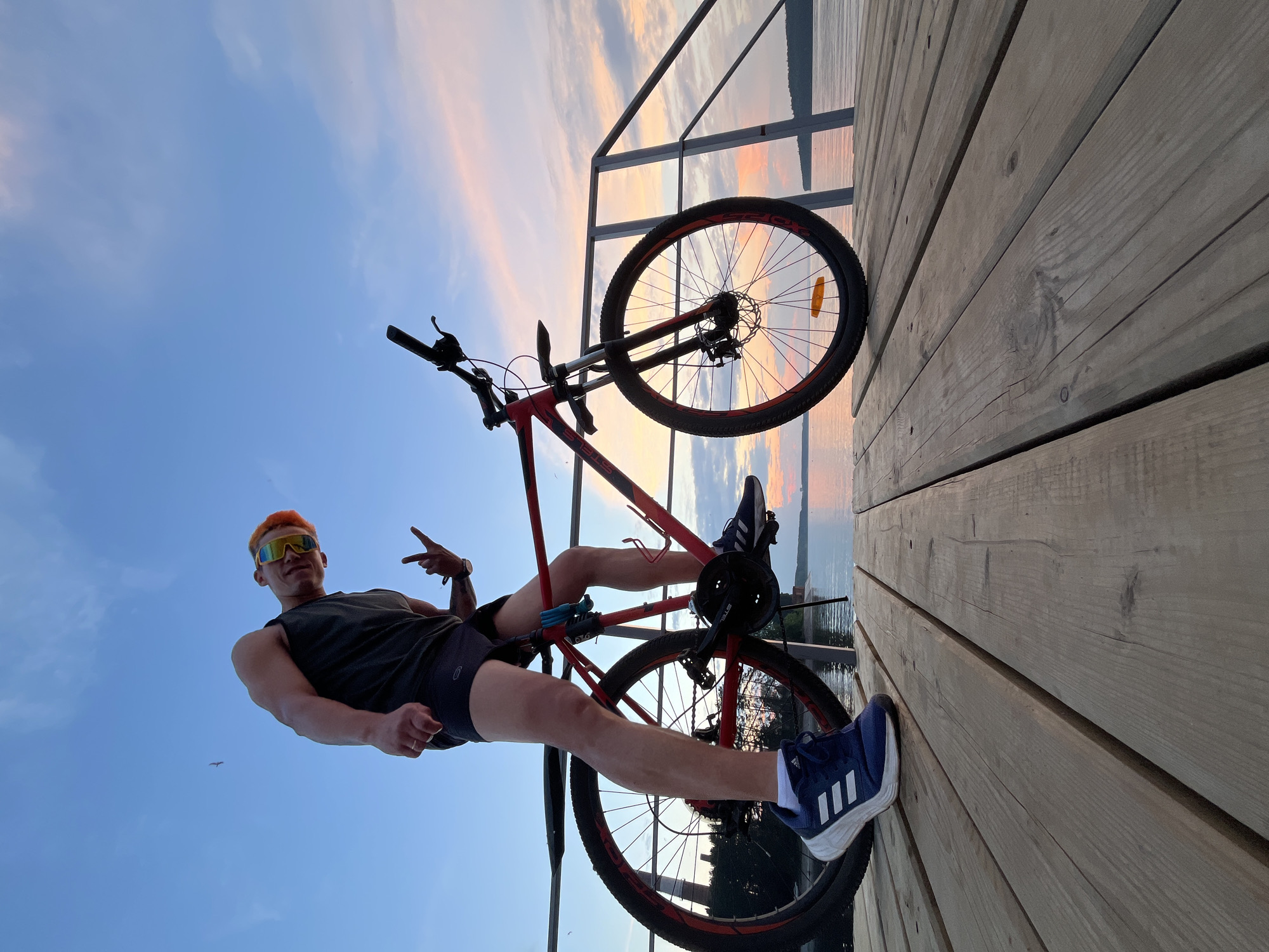 A young man with bright orange hair and sunglasses is posing with his bicycle on a wooden pier at sunset. He is wearing a dark sleeveless shirt, black shorts, and blue Adidas sneakers. The bicycle is red and black. The sky is a vibrant mix of blue, pink, and orange hues, indicating the setting sun over a body of water. In the background, trees and a distant structure are visible on the opposite shore. The pier is made of weathered wooden planks. The image captures a recreational moment by the water in Chaikovsky, Russia.