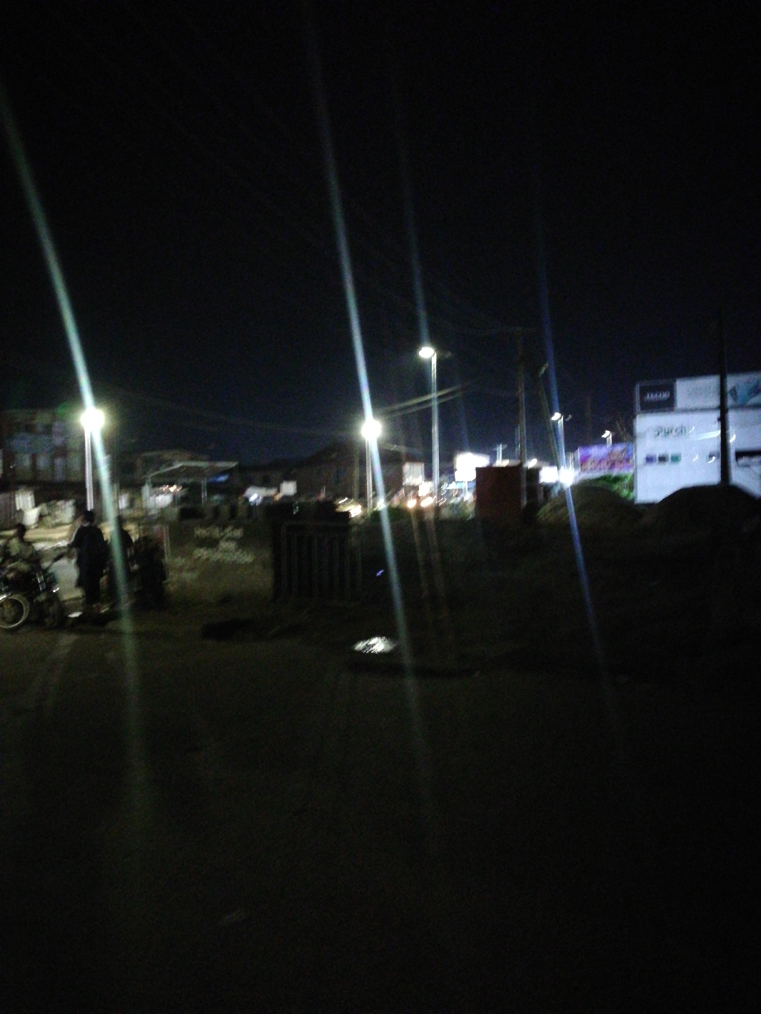 The image depicts a street scene at night in Lagos, Nigeria. The scene is dimly lit by streetlights, which cause prominent light streaks in the image. In the foreground, the street surface is visible. To the left, a group of people are standing near a motorcycle. Behind them, there is some kind of sign or wall. Further back, there are buildings and shops. A building with the word "purch" on it is visible in the background. The scene is somewhat blurred, and visibility is limited due to the darkness.