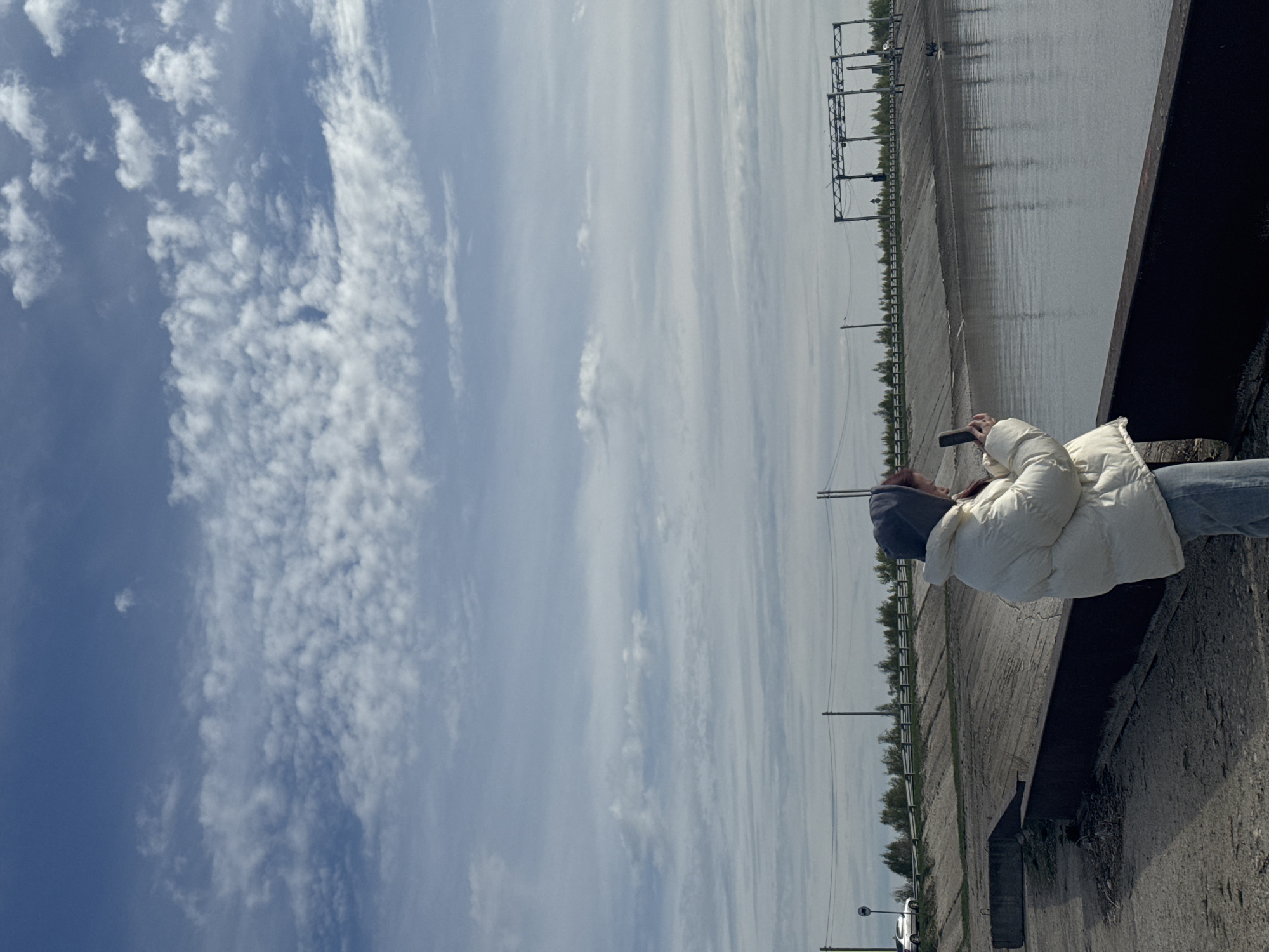 A photograph taken in Timiryazevskii S/S, Ivanovo, Russia shows a person leaning against a concrete barrier overlooking a reservoir or canal with a concrete bank. The person is wearing a white puffer jacket and a gray hooded sweatshirt. They appear to be holding a phone or camera. The water is still with some visible reflections. A metal grid structure runs along the reservoir bank, supported by metal poles. The sky is blue with patchy white clouds. The image is rotated, and the horizon is oriented vertically.