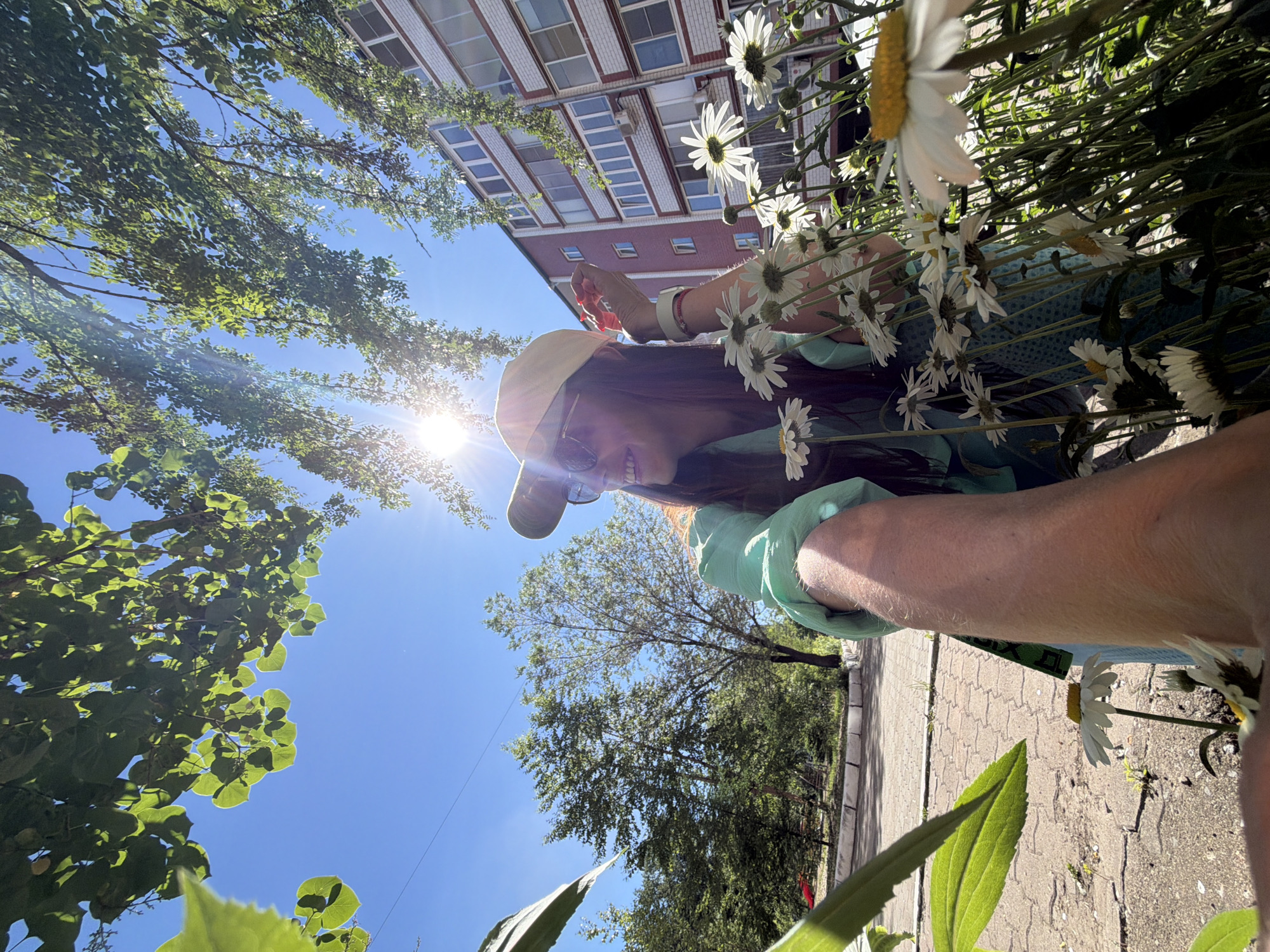 The image captures a young woman in a light green shirt and a tan baseball cap, posing for a selfie outdoors on a sunny day. She is smiling and wearing sunglasses, with a cluster of white daisies in the foreground, partially obscuring her. The background shows a modern apartment building with a red brick facade, trees, and a paved walkway. The overall scene suggests a casual moment in a residential area. The presence of a building with architecture typical of Russian apartment blocks, combined with trees and paving, points to a location in a city. While the provided context suggests Birobidzhan, Russia, the image itself does not contain explicit identifiers of this specific location. The lighting indicates daytime, with the sun casting strong shadows.