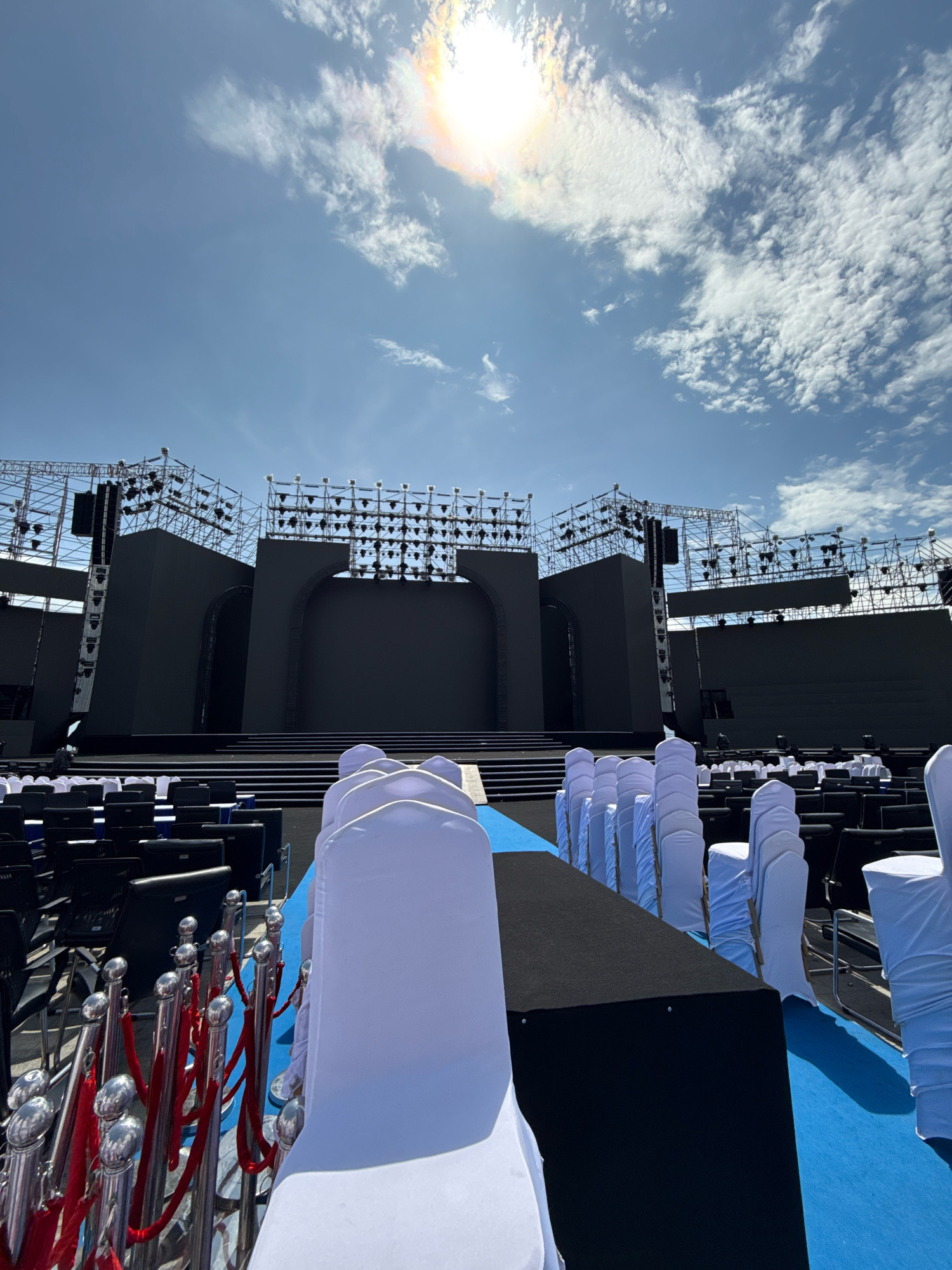 An outdoor event venue is set up with a large stage and seating area under a bright, sunny sky with scattered clouds. The stage consists of multiple black panels and is surrounded by scaffolding with numerous lights. Rows of black chairs are visible, some covered with white cloths, and a red rope barrier with stanchions is set up in the foreground. The scene suggests preparations for a concert or public event. Based on available information, this location is likely the Nha Trang Center in Nha Trang, Vietnam, a known venue for large-scale events.