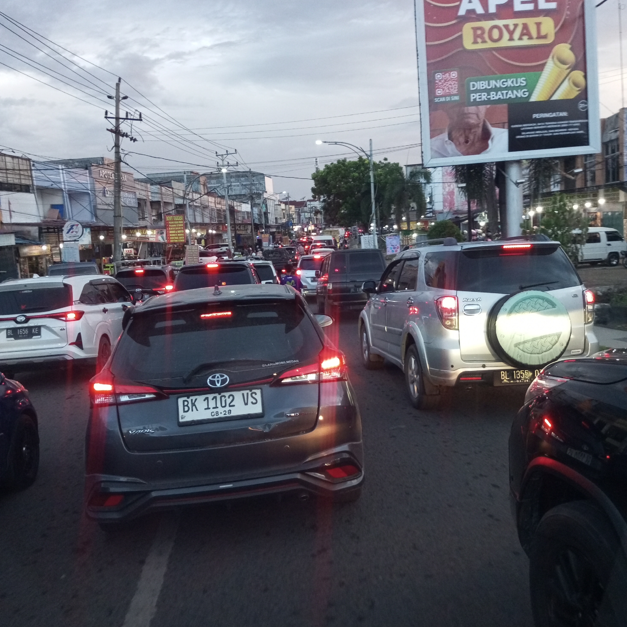 The image depicts a traffic jam in Lhokseumawe, Indonesia. Several cars are visible, including a gray Toyota Yaris with license plate BK 1102 VS and a silver Daihatsu Rush with license plate BL 1355 O. Various shops and buildings line the street, including a pharmacy ("APOTEK") and a store named "DONY VARIASI." A large "APEL ROYAL" advertisement is also visible. The sky is overcast.
