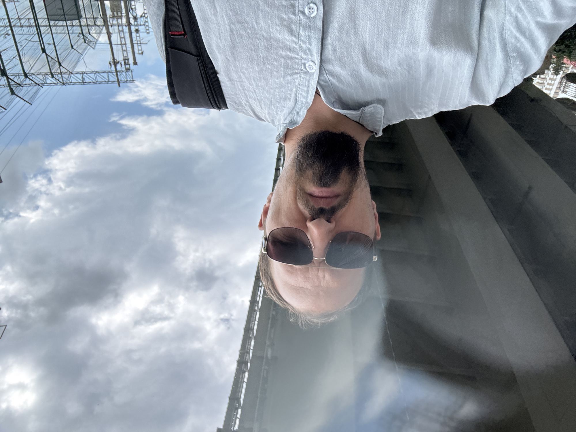 This is a rotated selfie of a man in Sochi, Russia. He is wearing a light blue button-down shirt and dark sunglasses. He has a beard and a goatee. In the background, there is a cloudy sky and what appears to be scaffolding or a construction site. The image is taken from a low angle, looking up at the man and the sky. The lighting suggests it is daytime, possibly overcast given the clouds.