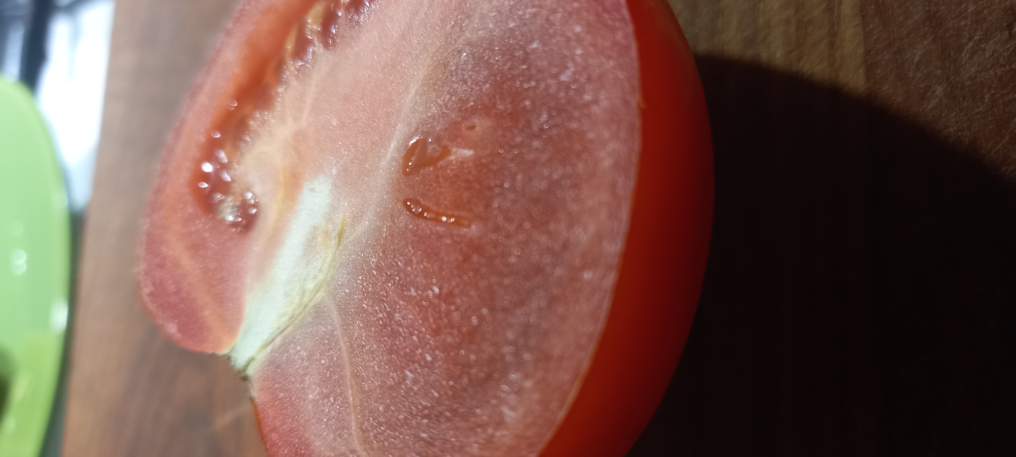 The image shows a close-up, diagonal view of a cut tomato on a wooden surface. The tomato is red and appears fresh, with visible seeds and pulp. A bright green object, possibly a plate or bowl, is blurred in the background. The wooden surface has a visible grain. There is no indication of the city of Kryvyi Rih, Ukraine, or any people or specific actions taking place.