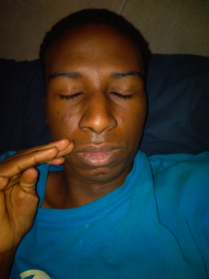 A close-up of a person with dark skin and short black hair, likely male, is seen with closed eyes. The individual is wearing a blue t-shirt and seems to be reclining against a dark blue pillow or headrest. Their right hand is resting near their cheek. The lighting is dim, casting shadows across their face, and the overall scene gives a calm or peaceful impression.
