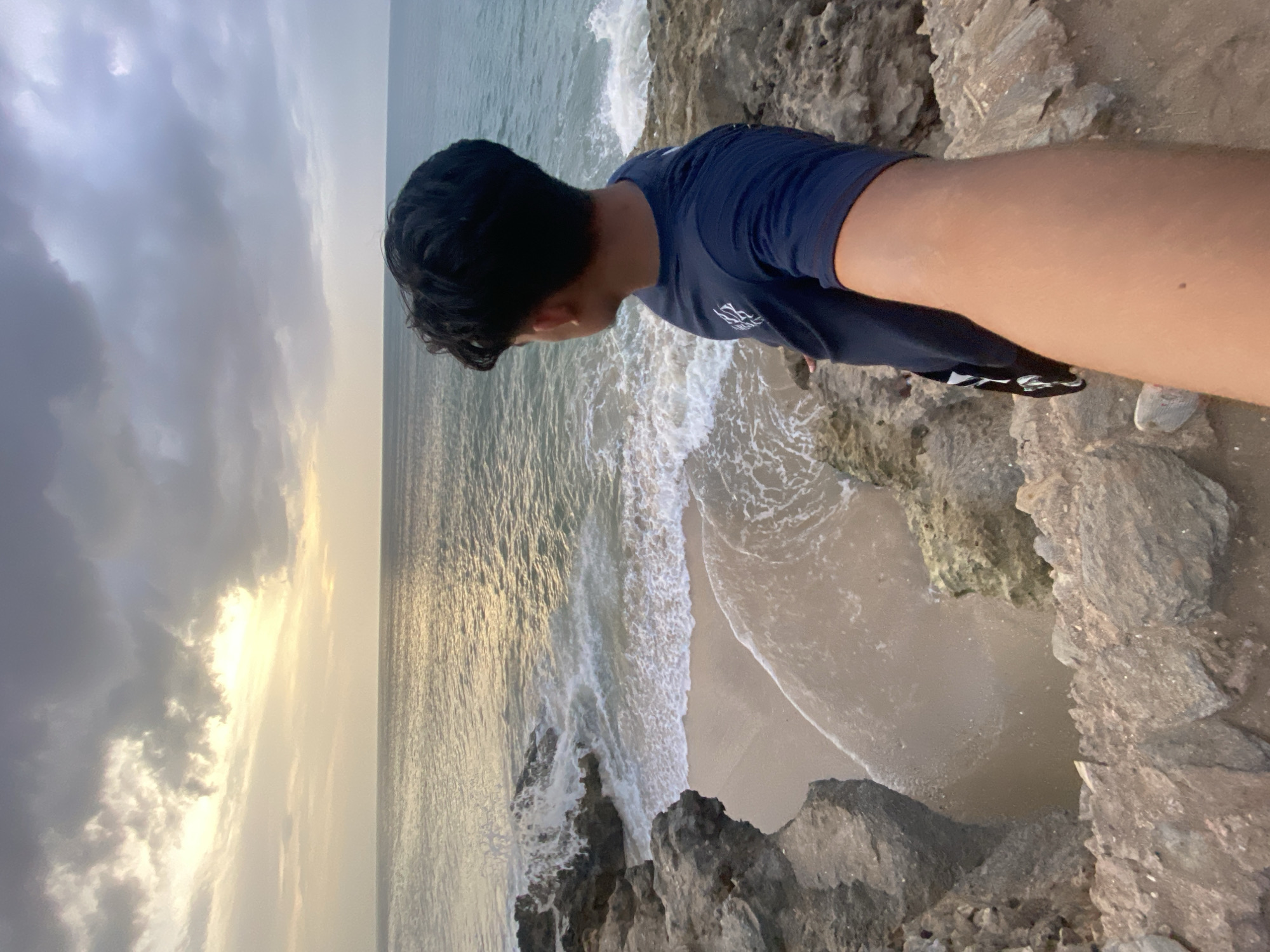 A person stands on a rocky cliff, looking down at the ocean. The waves are crashing against the rocks, and the beach is visible in the distance. The sky is cloudy, and the sun is setting. The location is Juan Griego, Venezuela. The person is wearing a blue t-shirt, shorts, and sneakers.