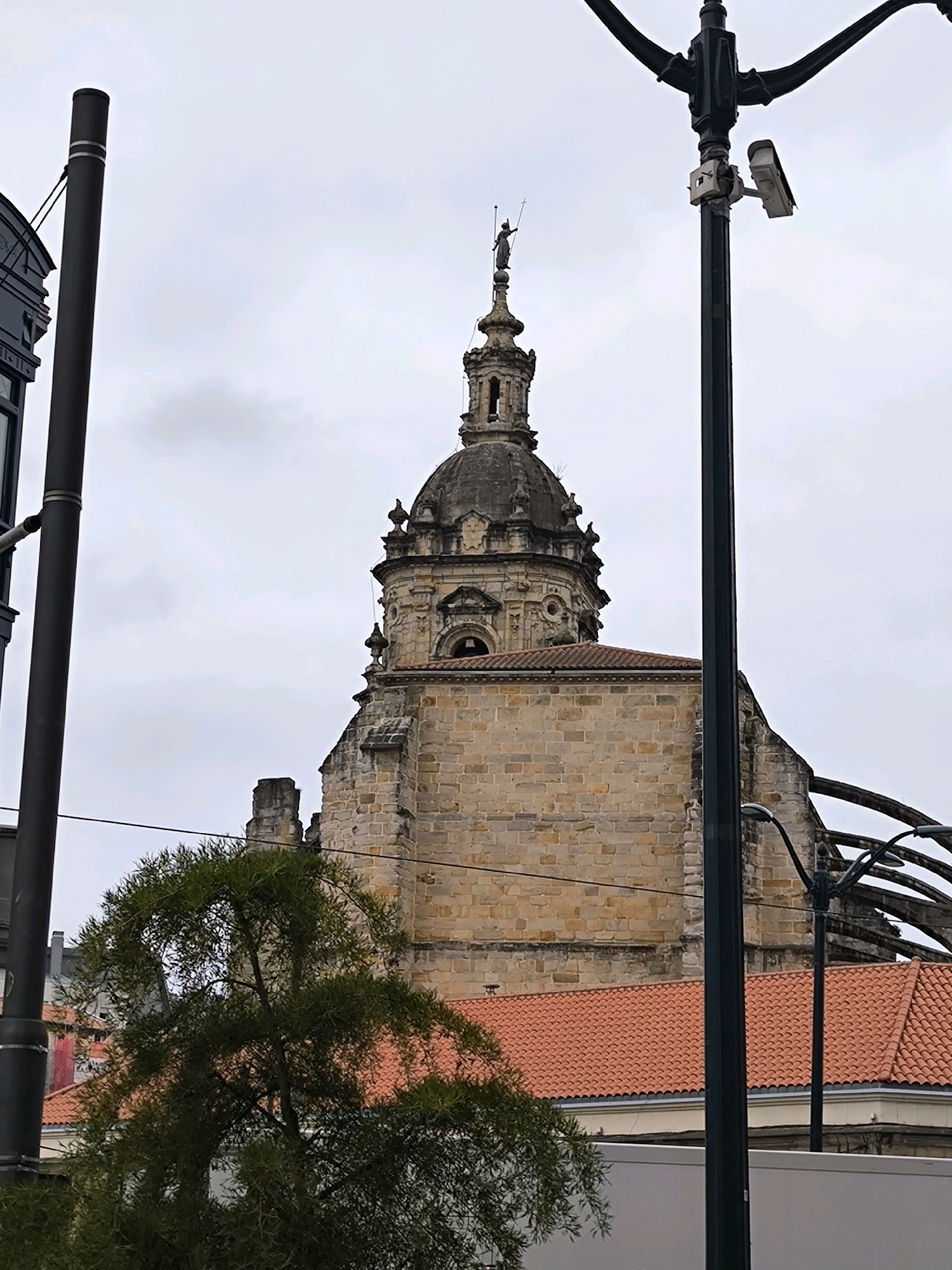 The image shows a view of the Bilbao Cathedral (Santiago Cathedral) in Bilbao, Spain, on an overcast day. In the foreground, two black lampposts with multiple arms are visible, one on the left and one on the right. The lamppost on the right has a white security camera mounted on it. A lush green tree with a distinctively curved shape stands in front of the cathedral. The cathedral's stone facade and a section of its red-tiled roof are visible. The sky is cloudy and light gray. The overall scene is a street-level perspective of an urban environment with a significant historical building.