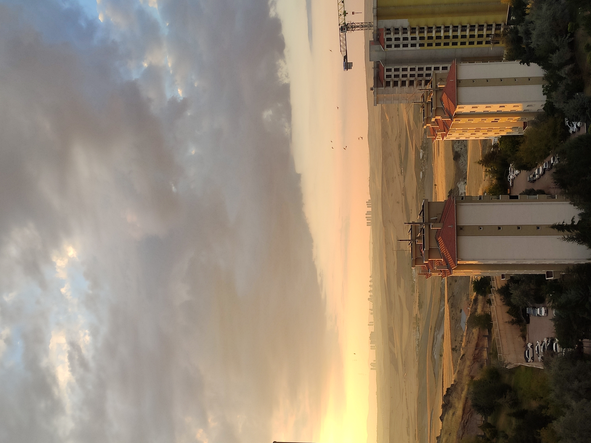 The image depicts a panoramic view of Etimesgut, Turkey, likely captured during the late afternoon or early evening given the warm, golden light. In the foreground, several multi-story residential buildings with red-tiled roofs are visible. Some of these buildings appear to be under construction, with unfinished facades and exposed windows. To the right, a large construction crane stands against the sky, indicating ongoing development in the area.
The middle ground is characterized by rolling hills and open, somewhat arid terrain, with patches of dry grass and sparse vegetation. A paved road or pathway winds through this landscape. In the distance, a city skyline is faintly visible, characterized by numerous high-rise buildings silhouetted against the horizon.
The sky is dramatic, filled with a mix of dark, heavy clouds and patches of lighter, illuminated sky, suggesting a possible change in weather or the aftermath of rain. A few small birds are visible in flight against the sky. A parking lot with several cars is situated near the foreground buildings, suggesting residential activity. The overall impression is one of a developing urban fringe transitioning into a more natural, hilly landscape.