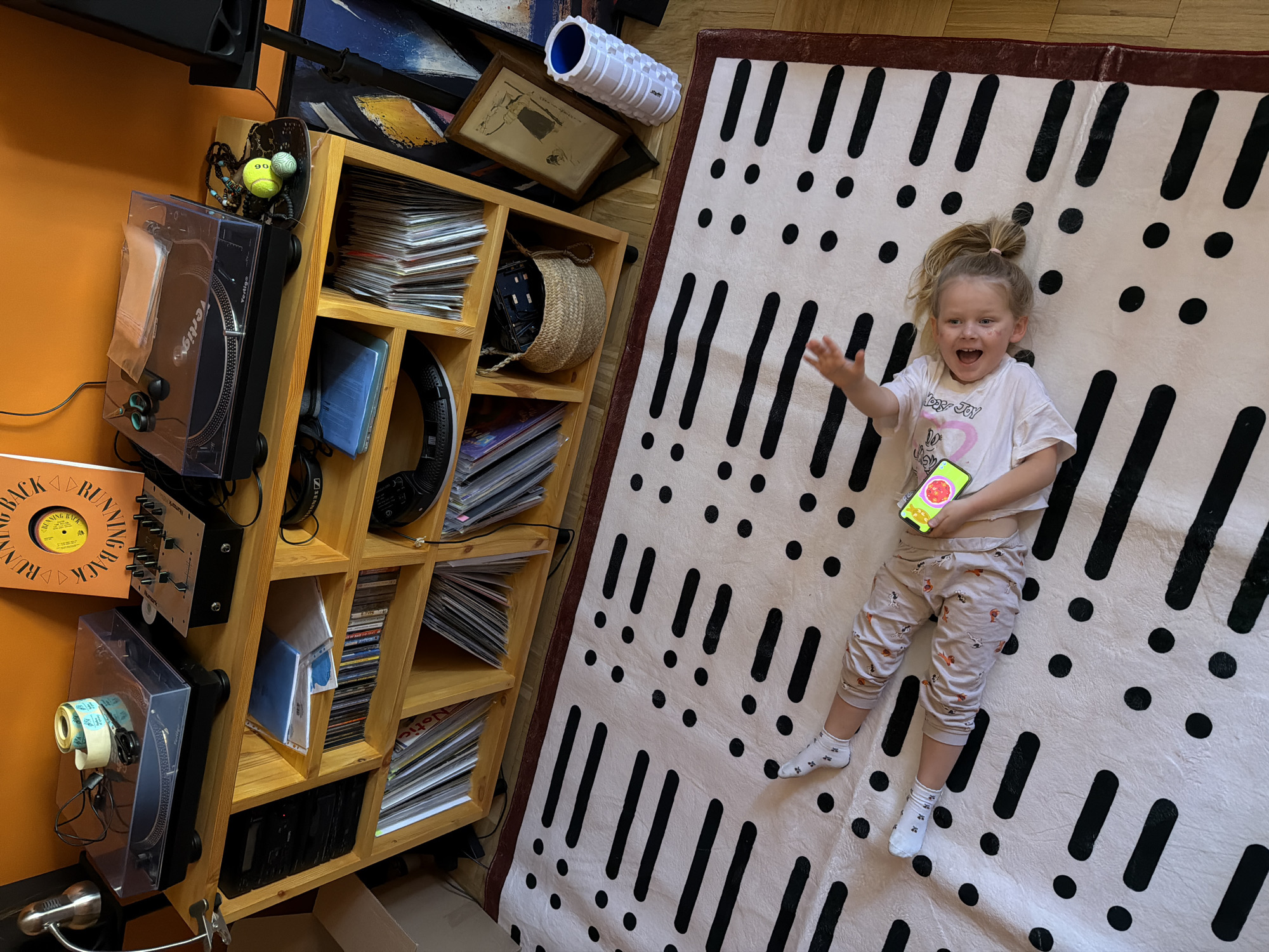 This indoor scene, likely set in a home in Khimki, Russia, features a delighted young child lying on a large, cream-colored rug. The rug has a distinctive pattern of black dots and elongated vertical dashes, resembling exclamation marks. The child, a toddler with light-colored hair pulled into a high, messy bun, smiles widely with an open mouth and outstretched right arm, conveying clear excitement or joy. They are dressed in a white t-shirt with visible text including "Peace Joy Love" and "I <3 My" (partially obscured), light-colored patterned pajama pants adorned with small figures, and white socks. In their left hand, they hold a smartphone displaying an app with a bright red, tomato-like icon on a green and yellow background, alongside some numerical display.
Adjacent to the rug, a tall, light-wood shelving unit is packed with a variety of items, set against a vibrant orange wall. The shelves are predominantly filled with stacks of vinyl records. Two turntables are visible, one mounted to the side of the unit with a small roll of tape and scissors resting on its dust cover, and another below it. A pair of black Sennheiser headphones hangs from a shelf, and a silver audio mixer is mounted above one turntable. Other objects on or around the shelving unit include framed artworks, a white foam roller with "Trigger Point" visible, a woven basket, and two tennis balls. An orange record sleeve with "RUNNING BACK" and "YELLOW BACK" text in a circular design is propped up near the base of the unit. A small metallic lamp is also visible at the bottom left. The scene captures a moment of playful domestic life.