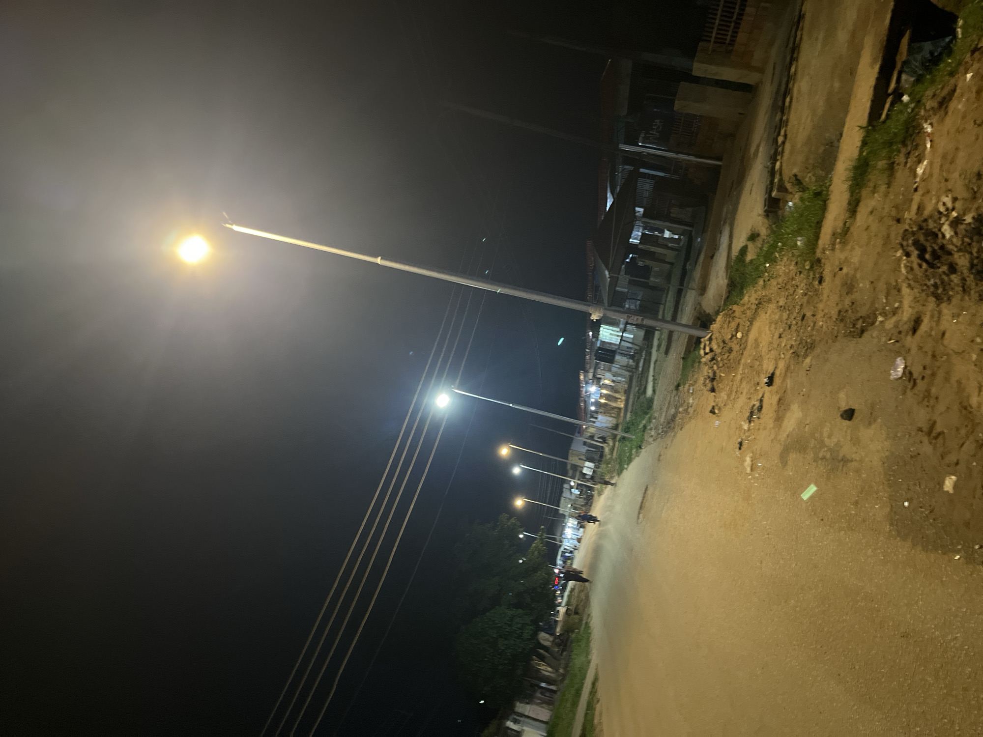 The image depicts a street scene at night in Abuja, Nigeria, illuminated by streetlights. The road is unpaved and appears to be made of dirt or gravel. Several streetlights are visible, casting a yellow glow on the surroundings. Buildings, some with corrugated metal roofs, line the street on the right side. There are also trees and power lines visible. A few people are walking along the road, and at least one motorcycle is present. The ground is littered with debris, suggesting an unkempt environment. The overall impression is of a residential or commercial area at night in a developing part of the city.