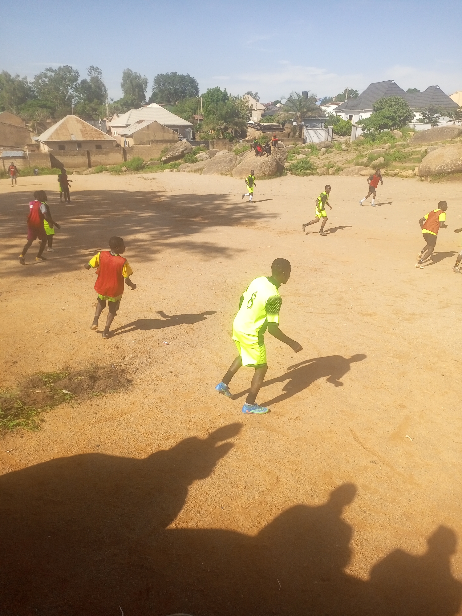 This is a medium-angle, eye-level shot of a group of young boys playing soccer on a dirt field in Jos, Nigeria. The sun is shining brightly, casting long shadows across the sandy ground. In the background, there are several buildings with corrugated metal roofs, typical of residential areas in Jos. Some large rocks and sparse vegetation are also visible.
The boys are wearing various colorful jerseys and shorts, indicating they are engaged in a casual game. One boy in the foreground is wearing a bright yellow and green soccer uniform and appears to be dribbling the ball. Another boy in a red and yellow uniform is running towards the left of the frame. Other boys are scattered across the field, some running, some watching. The overall atmosphere is one of energetic outdoor activity.
