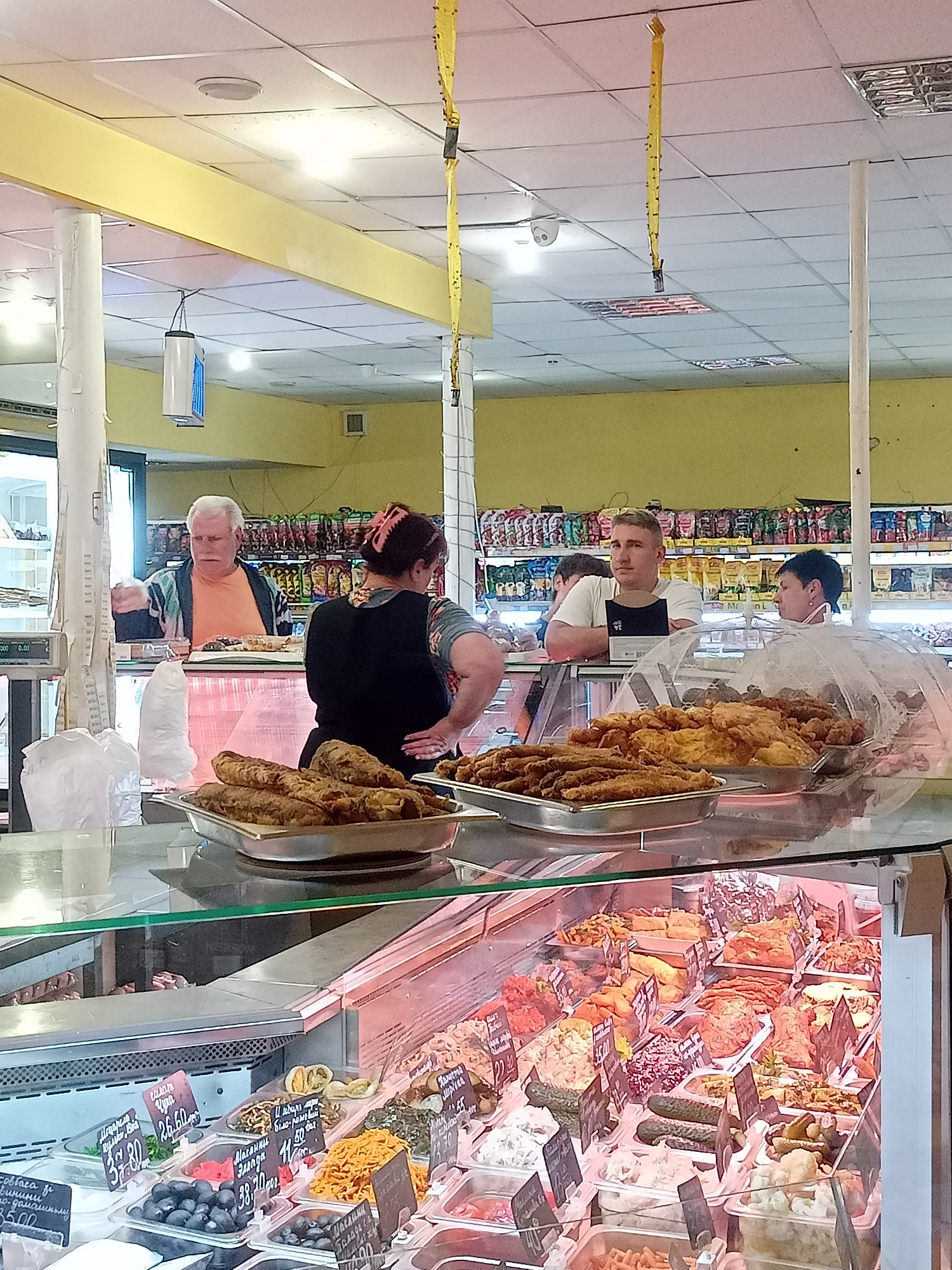 The image depicts a market stall in Bilhorod-Dnistrovskyi, Ukraine, featuring a variety of prepared foods displayed in refrigerated cases. In the foreground, several trays of fried food, possibly fish or chicken, are visible. Behind them, the refrigerated display case contains numerous salads and pickled items. Three individuals are present behind the counter. An older man with white hair, wearing an orange shirt and a patterned cardigan, is interacting with a customer. To his right, a woman with dark hair in a bun, wearing a black apron over a patterned shirt, stands with her hands on her hips. Further to the right, a young man in a white t-shirt and dark apron is looking towards the camera. In the background, shelves stocked with various food products are visible. The environment suggests a food retail setting with bright lighting and clean displays.