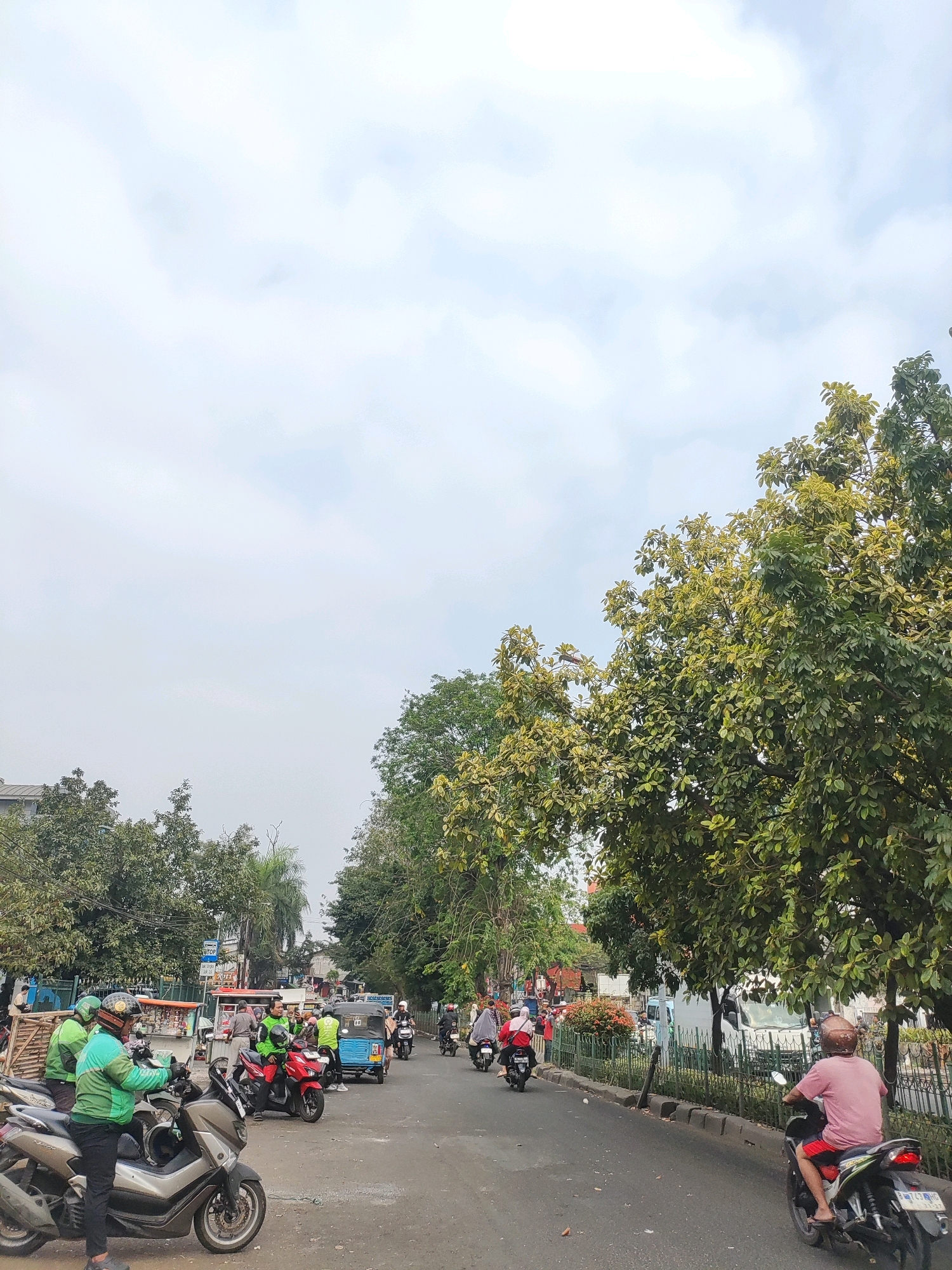 The image depicts a street scene in East Jakarta, Indonesia, characterized by moderate traffic and lush greenery. In the foreground, several motorcycles are parked, with some individuals wearing green jackets, likely motorcycle taxi drivers, standing near them. One person in a pink shirt and red shorts is riding a motorcycle away from the camera on the right side of the road. A blue motorized rickshaw, also known as a "bajaj," is visible in the middle of the road, suggesting a common mode of transportation in the area. Pedestrians and other vehicles are present further down the street, indicating a bustling urban environment. The background features buildings and a variety of trees, their foliage creating a canopy that partially obscures the overcast sky. The road surface appears to be asphalt, with some debris scattered along the edges. The overall atmosphere is that of a typical day in a busy Indonesian city.