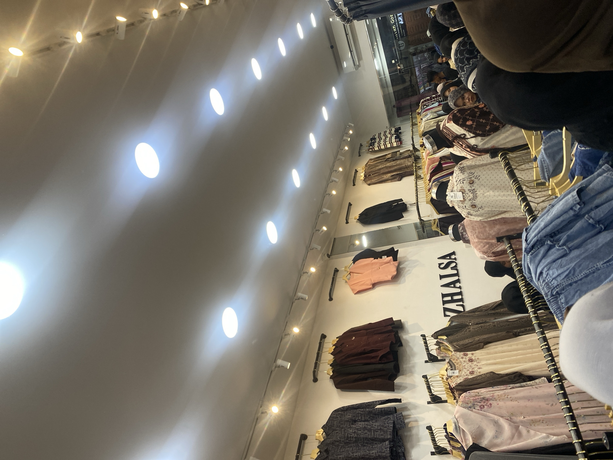 The image shows an interior view of a clothing store, presumably in Lhokseumawe, Indonesia, based on the prompt. The store features rows of clothing displayed on racks, including shirts and jackets. The clothing hangs on hangers attached to the wall. A sign with the name "ZHALSA" is visible on the wall, likely the store's name. People are browsing and looking at clothes. Fluorescent lights illuminate the area.