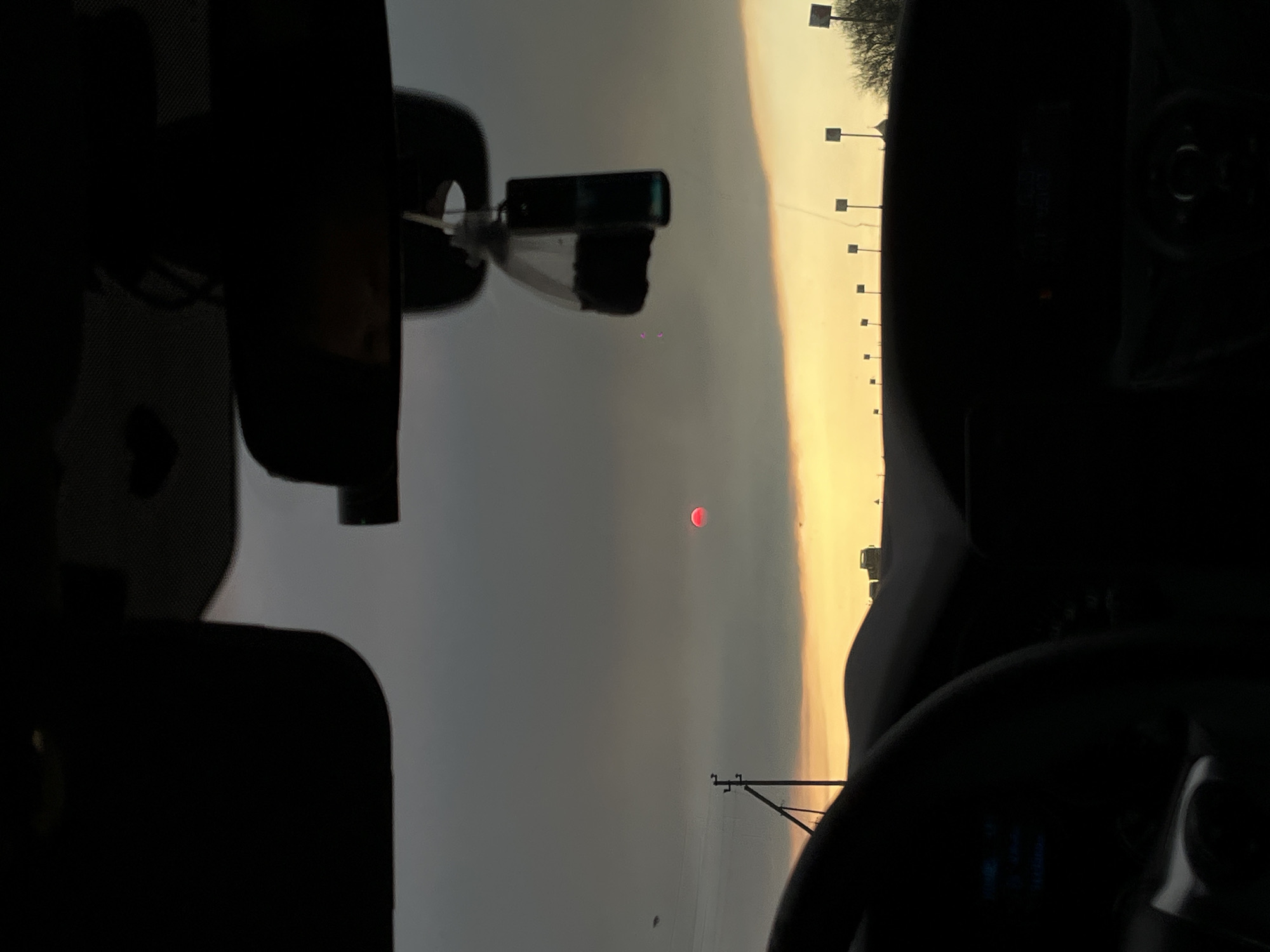 The image captures a scene from inside a vehicle, looking out through the windshield at a hazy sunset or sunrise in Beysug, Russia. The sky is divided into a vibrant golden-orange band along the horizon on the right, transitioning to a duller, grey-blue hue across the rest of the frame. A distinct, deep red-orange sun, appearing as a disc, is prominently positioned in the upper-left sky, partially obscured by atmospheric haze.

The foreground is dominated by the dark silhouettes of the car's interior. On the left, the dashboard and a portion of the steering wheel are visible, with faint, unreadable blue digital text illuminated on the instrument cluster. On the right, the rear-view mirror is visible, showing a dark reflection, and what appears to be a phone holder or similar device is attached to it. Along the distant horizon, a series of silhouetted utility poles or streetlights with square tops are regularly spaced, and some bare trees are visible on the far right. The scene conveys a sense of tranquility, observed during travel or a stop, under hazy atmospheric conditions at either dawn or dusk.