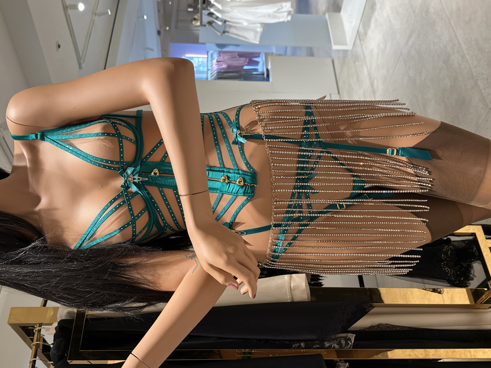 A full-length, medium shot shows a mannequin wearing a strappy, teal lingerie set with rhinestone accents, and hose in a retail setting. The mannequin is dark-haired and has light brown skin, standing in front of clothing racks. The lingerie set is elaborate and features a chained fringe detail at the hips. The environment appears to be a clothing store, based in London, UK.