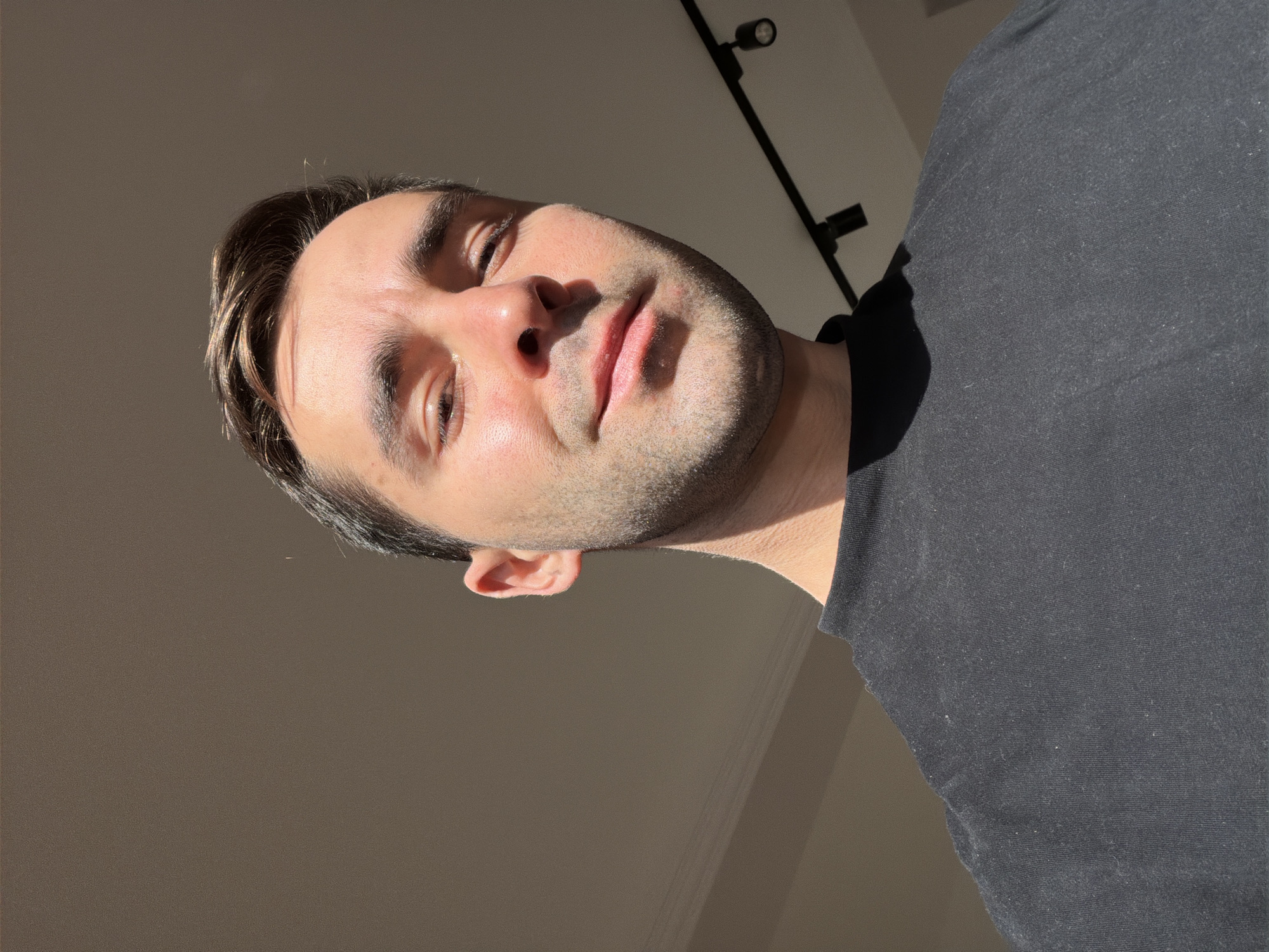 A close-up, low-angle portrait captures a fair-skinned man with short dark hair and stubble. He wears a dark-colored t-shirt, and the lighting creates shadows on his face. The background is a neutral-toned wall, with a black track lighting fixture visible in the upper right corner. There is no indication of the location being Tbilisi, Georgia.