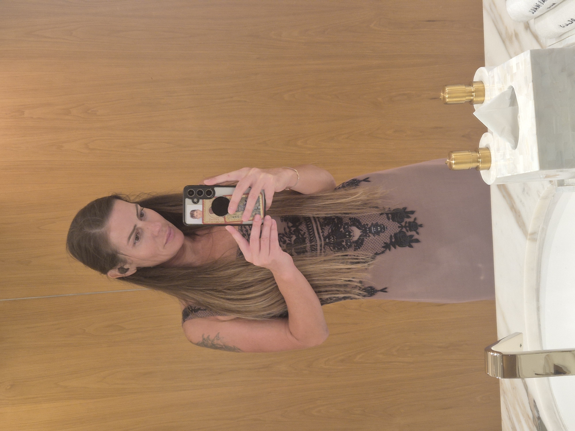 The image, correctly oriented, features a woman taking a mirror selfie in what appears to be a bathroom or dressing room. She has long, straight brown hair and is wearing a light-colored dress or top with black lace detailing on the shoulders and chest area. She holds a dark-colored smartphone in her right hand, aiming it towards the mirror to capture her reflection. The phone case is personalized with a small image of a person.
Notable details include a thin bracelet on her left wrist and a dark tattoo visible on her left forearm. A black earbud is also present in her left ear. The background behind her reflection is a warm, light brown wooden wall or paneling.
On the white countertop to the right of her reflection, several items are visible: a square tissue box dispenser crafted from a luminous material resembling mother-of-pearl or shell, adorned with two gold-colored decorative finials. Adjacent to it are stacked white towels, one of which clearly displays embroidered Arabic script. A shiny, gold-colored towel bar or handle is also partially visible on the far right. The overall setting suggests an interior space, likely well-appointed, possibly in a hotel or a private residence. The woman maintains a neutral expression, looking directly towards the phone camera in the reflection.