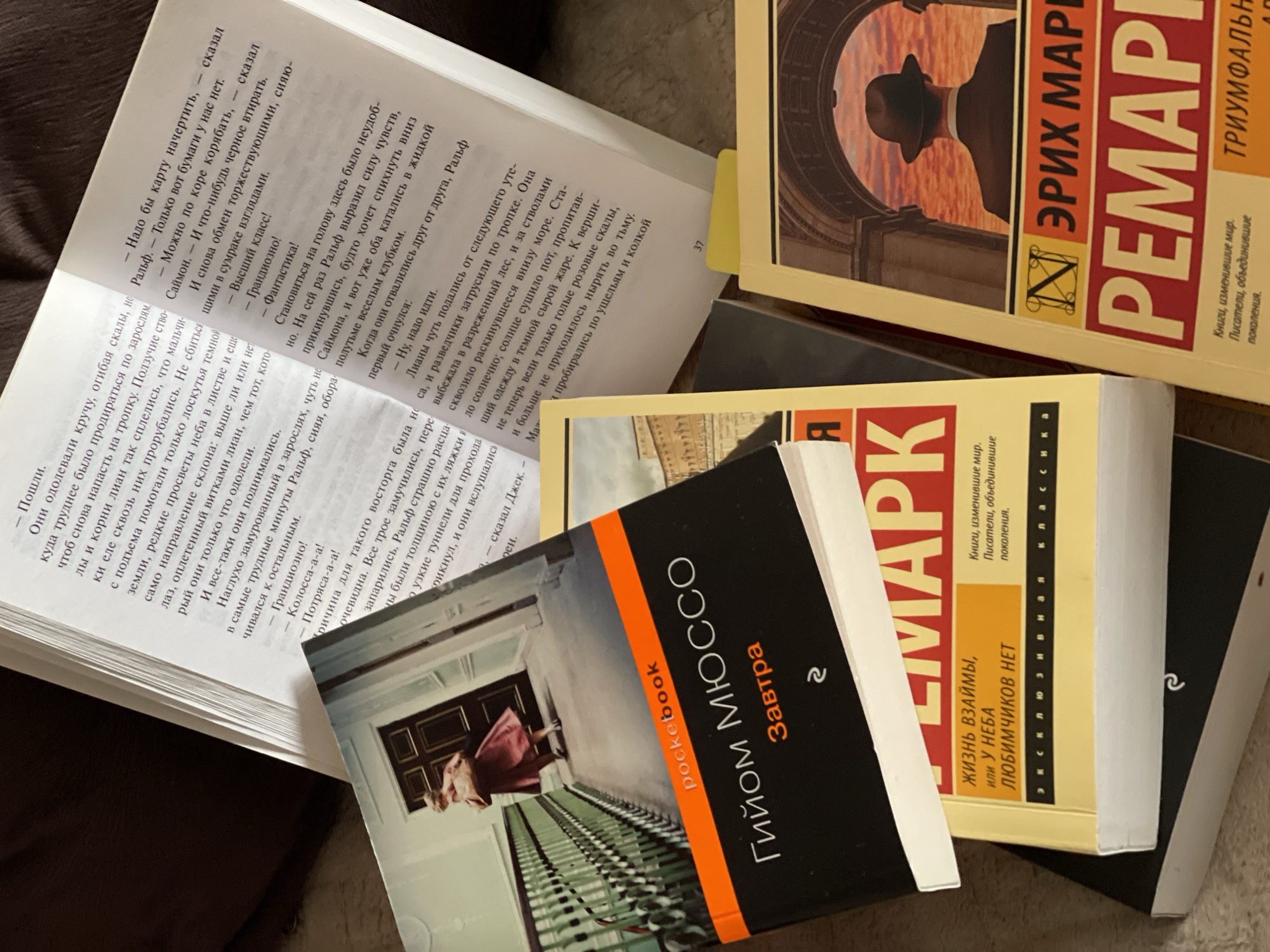 The image shows a close-up, overhead view of several books stacked and lying open on a textured surface, possibly a table or bed. The books are primarily in Russian. One prominently featured book is titled "Завтра" by Гийом Мюссо, with "pocket book" printed above the title. Another book, yellow with red text, displays "ЖИЗНЬ ВЗАИМЫ, ИЛИ У НЕБА ЛЮБИМЧИКОВ НЕТ" and "ЭКСКЛЮЗИВНАЯ КЛАССИКА". A third book, also with red text, shows "ЭРИХ МАРИ РЕМА". The open pages of the books reveal lines of text. The lighting is natural and appears to be coming from the top left. The scene is static, with no active subjects or interactions. There is no identifiable location information, city, or country mentioned or depicted in the image.