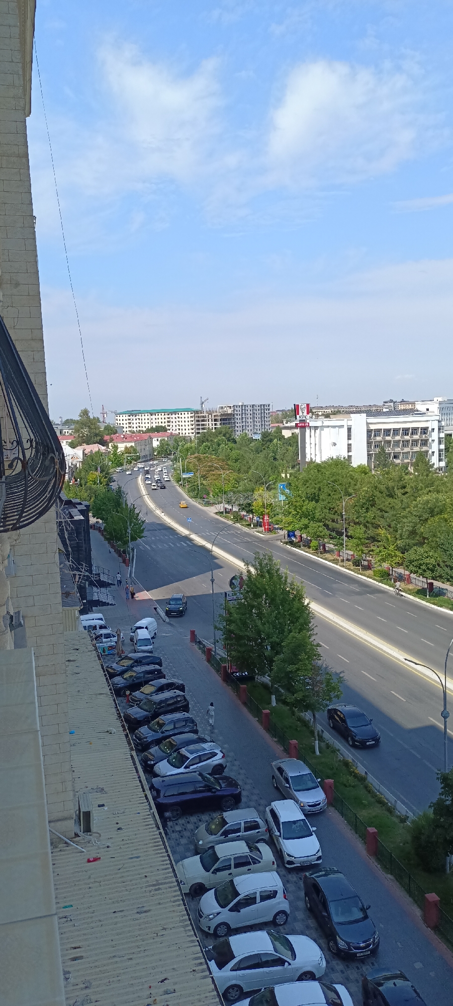 The image captures a street scene in Fergana City, Uzbekistan, with a focus on a multi-lane road lined with trees and buildings. The foreground shows a parking area filled with numerous cars, predominantly sedans and SUVs, of various colors including black, white, and silver. A few individuals are visible walking in the parking area and on the sidewalk adjacent to the street. The road itself is bustling with traffic, featuring cars and at least one yellow taxi. Beyond the road, modern buildings, including an apartment complex and commercial structures, are interspersed with lush green trees, indicating a developed urban environment. One building prominently displays a KFC logo. The sky above is a clear blue with scattered white clouds, suggesting fair weather. The perspective is from an elevated position, likely a balcony or window, looking down and across the street. The overall impression is a dynamic urban landscape on a sunny day.