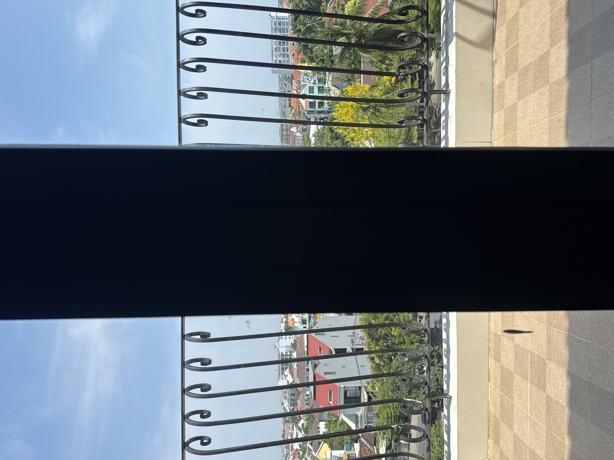 The image depicts a view from a balcony, likely in Hội An Ward, Vietnam, based on visual characteristics. The foreground features a decorative wrought-iron railing partially obscuring the cityscape below. The railing is painted a dark color. A dark, horizontally-oriented, partially visible object occupies the center of the frame, possibly a portion of the balcony structure or a window frame. Beyond the railing, the cityscape includes buildings of varying heights and colors, interspersed with trees and vegetation. The sky is light blue. The balcony floor appears to be tiled. The lighting suggests it is daytime.