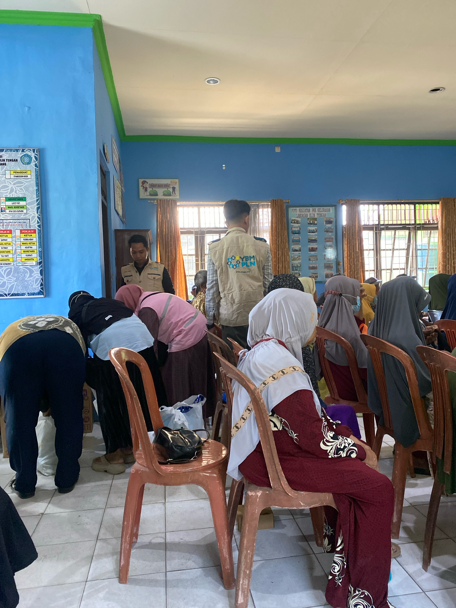 The image depicts an indoor gathering in what appears to be a community hall or office in Banjarbaru, Indonesia. The room has bright blue walls with a green trim along the ceiling. Several individuals are present, most of whom are women wearing hijabs and modest attire, some seated and others standing and bending over. One man in a tan vest with "YBM PLN" printed on it is standing and facing away from the camera. Another man, possibly an organizer, is standing and looking down at the people gathered. The floor is tiled, and the room is furnished with simple plastic chairs and wooden cabinets. There are posters and framed pictures on the walls. The scene suggests a meeting, a distribution event, or a community program.