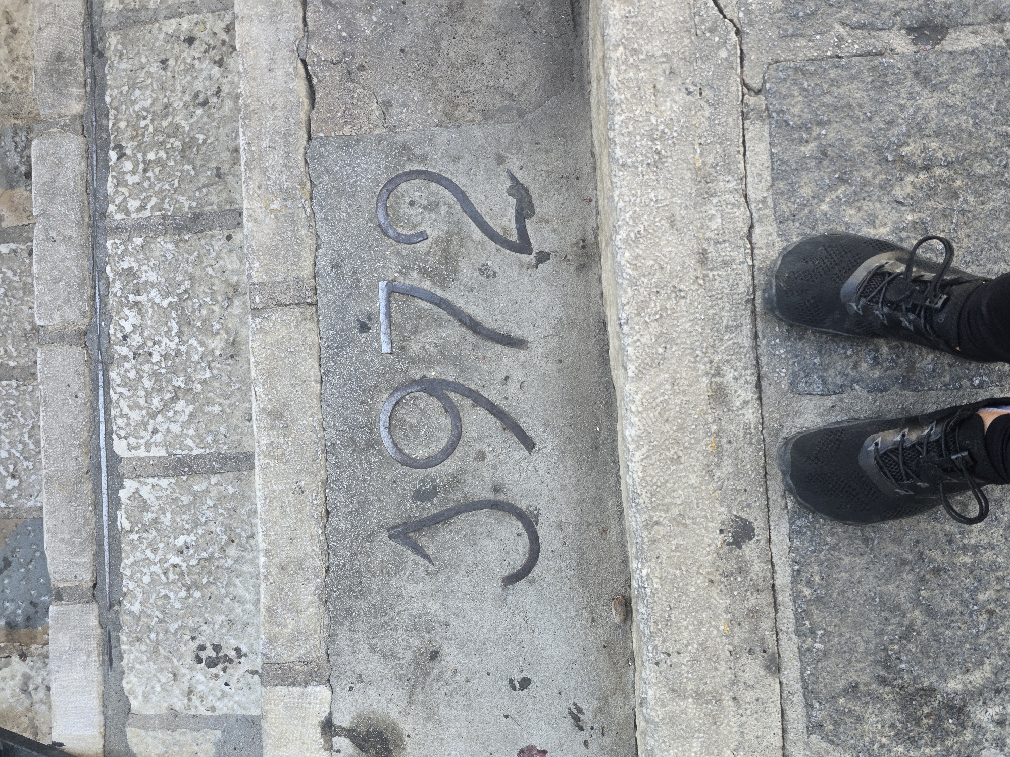 The image shows a close-up view of the number "1972" embedded into concrete steps. The numerals are dark and metallic, appearing to be made of wrought iron or a similar material. The steps themselves are constructed from rough, grey stone blocks, showing signs of age and wear. In the lower portion of the frame, the black athletic shoes of a person are visible, standing on the lower steps. This appears to be a public walkway, possibly in the town of Mijas, Spain, where such historical markers are common.
