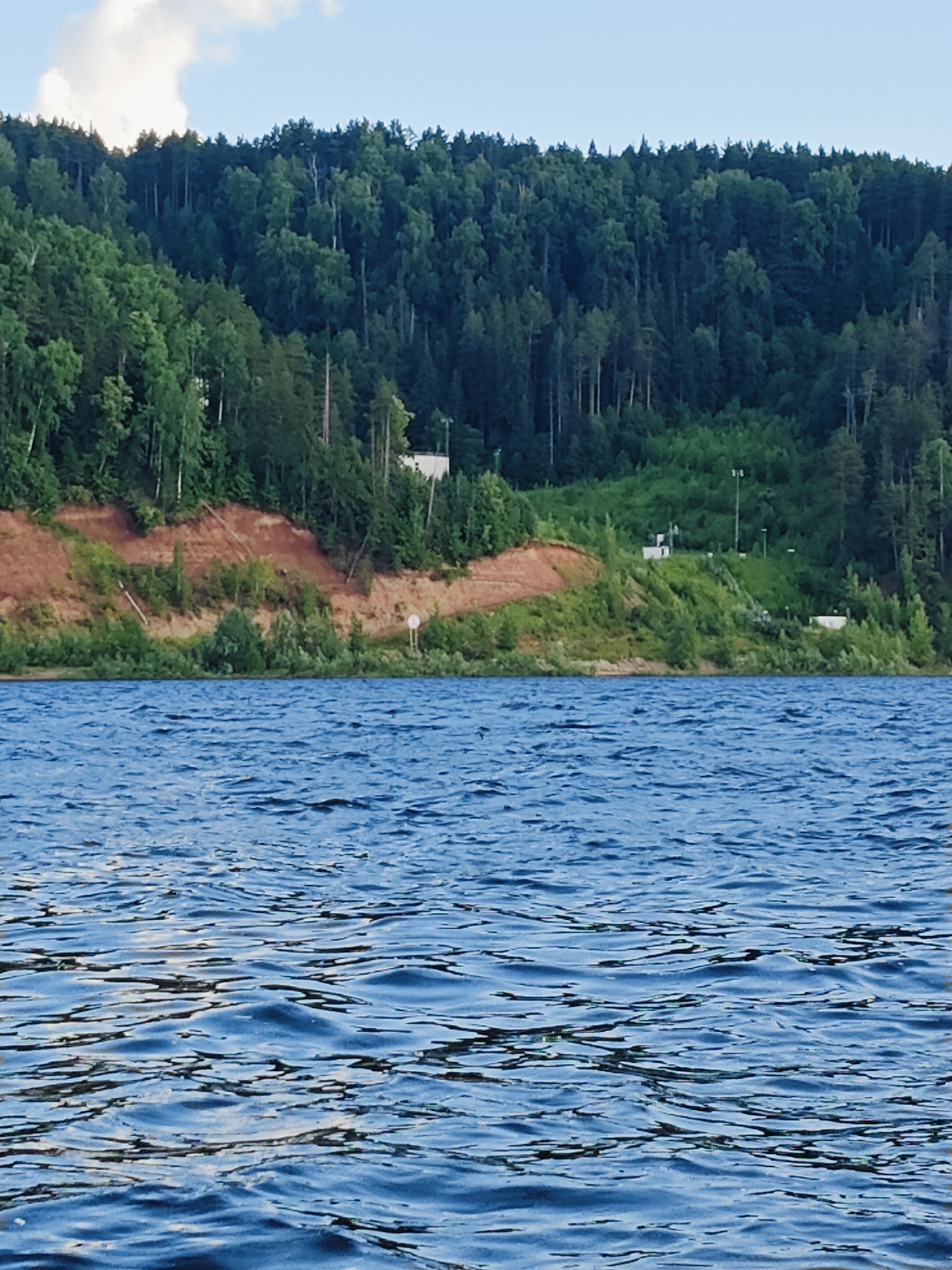The image shows a body of water in the foreground with ripples on its surface, reflecting the sky and surrounding landscape. In the midground, a steep, reddish-brown bank rises, partially covered by green vegetation and trees. A dense forest covers the upper part of the bank and extends to the horizon. Scattered structures, possibly buildings or infrastructure, are visible on the green slopes. The sky is partly cloudy with patches of blue. The location is identified as being in the city of Prikamsky, Russia.