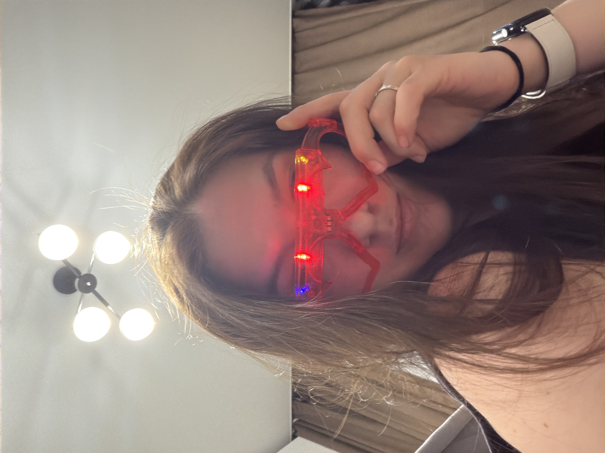 A fair-skinned woman with shoulder-length brown hair is positioned with her head tilted to the right. She's wearing red, light-up glasses that appear to be resting on her forehead rather than being worn in the traditional manner. On her left wrist, she wears a smartwatch with a white band and a black band underneath. A ring is visible on her left ring finger. Behind her to the left is a white wall with a modern light fixture featuring multiple spherical lights. To her right is what appears to be a sofa or bed with neutral-colored upholstery. The location, Staraya, Russia is not discernible from this image alone. The overall composition suggests a casual selfie taken indoors.