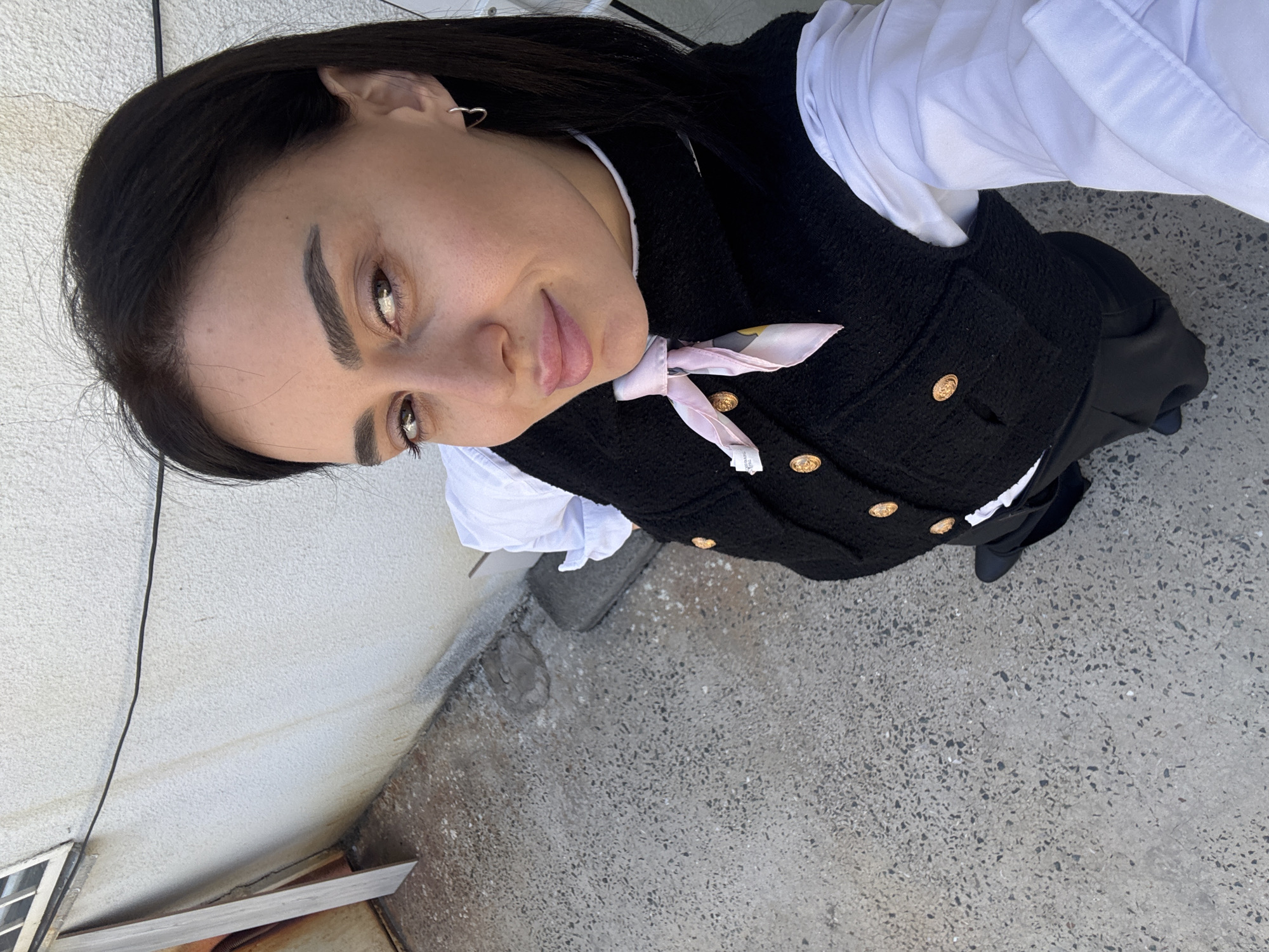 An outdoor, low-angle shot captures a young woman looking down directly at the camera with a subtle smile. The image is rotated, giving it a dynamic perspective. She has dark hair, dark eyes, and fair skin, with her eyebrows and eyes visibly defined with makeup.
The woman is dressed in a white long-sleeved shirt layered under a black, textured vest with prominent gold-toned buttons. A light pink scarf, adorned with a delicate pattern, is tied neatly around her neck. A silver, heart-shaped earring is visible in her left ear. Her lower attire includes dark trousers or a skirt, paired with black shoes.
The background features a light-colored wall with a rough, stucco-like texture, displaying some minor discolouration and stains. A dark cable runs vertically along the wall. The ground is a light grey concrete surface, typical of urban environments, with scattered dark aggregate specks. Some wooden planks or debris are visible near the base of the wall. The lighting indicates it is daytime. A small label with illegible text can be seen attached to her vest or scarf. This scene is set in Tbilisi, Georgia.