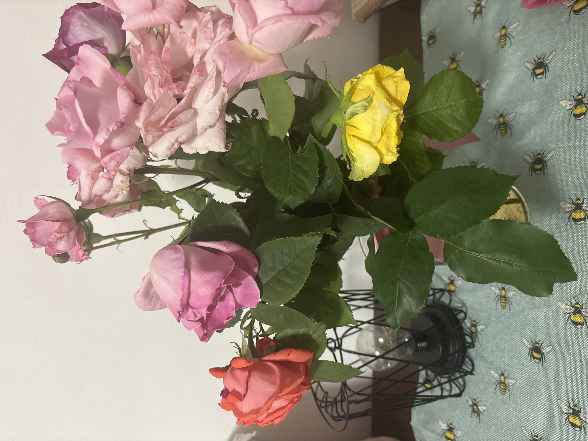 A bouquet of roses is featured prominently, presented in a decorative black wire cage that resembles a lamp base or a modern vase. The roses display a range of colors, predominantly various shades of pink, from light and delicate to deeper, more saturated tones, with some petals showing hints of purple. Many of the pink roses appear to be past their prime, with noticeable wilting, creased petals, and some browning edges. In stark contrast, a vibrant yellow rose stands out on the right side of the arrangement, and a coral-orange rose is visible towards the bottom, both appearing relatively fresher. Abundant green leaves intersperse throughout the bouquet.
The setting appears to be indoors. The background on the left is a plain white surface, likely a wall. On the right, a light blue fabric with a playful pattern of small yellow and black bumblebees serves as a tablecloth or mat. No specific activity is taking place; the image is a still life. There are no people visible, nor any discernible text. The lighting is soft and consistent, indicative of an interior environment.