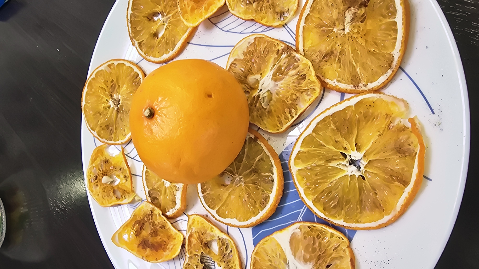 A high-angle, close-up photograph displays an arrangement of fruit on a white, round plate featuring a minimalist blue line pattern. The central object is one whole, unpeeled orange, positioned with its stem-end visible at the top, exhibiting a smooth, bright orange peel with a minor dark indentation on its side. Surrounding the whole orange are approximately twenty-two slices of what appear to be dried or dehydrated oranges, varying in size and shape. Many slices are circular cross-sections, revealing the shrunken, translucent yellow-orange pulp and internal segmentation. Other slices are irregular, broken, or partial segments, some exhibiting darkened or browned areas consistent with dehydration or light cooking. Small black particulates, consistent with ground spice, are scattered sparsely across several orange slices and the white surface of the plate. A few visible seeds are present within the pulp of some slices. The plate rests on a dark, non-reflective surface, likely a countertop or table, which occupies the background and shows subtle, faint reflections in the upper portion of the frame. No individuals are present, and no active interactions are depicted. The scene is located in Kazan, Russia.