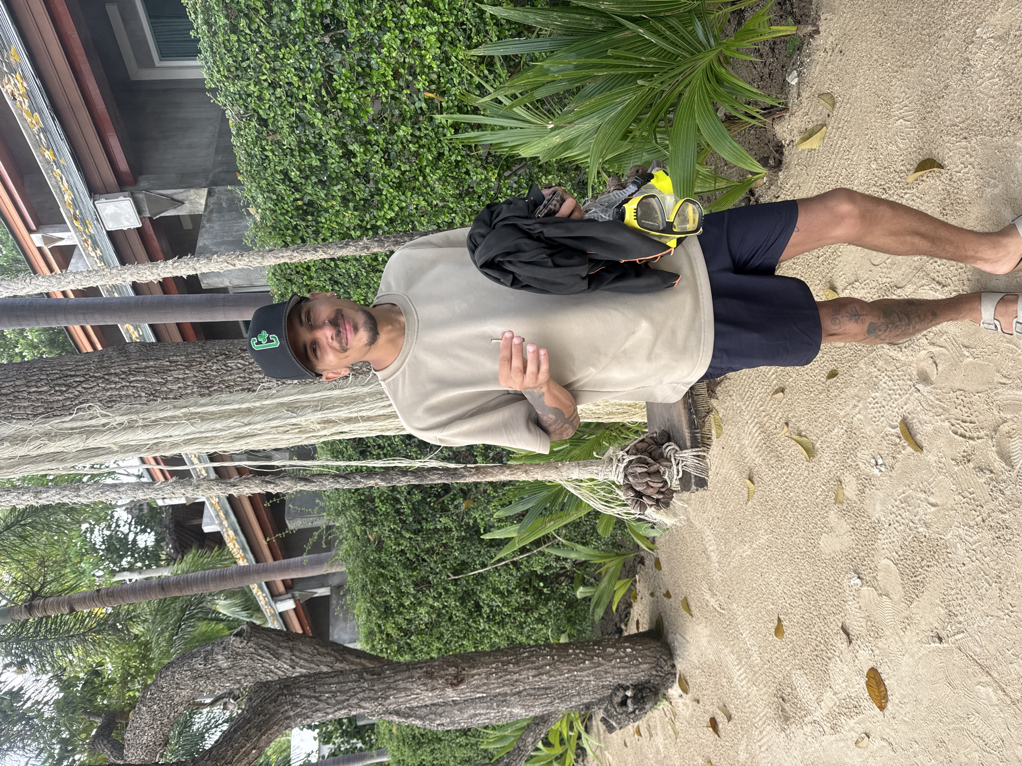 A man in a black cap, beige shirt, and blue shorts stands on a sandy surface in ตำบลเกาะพะงัน, Thailand. He holds black clothing and yellow goggles. A tree and greenery are behind him, along with ropes and a building.