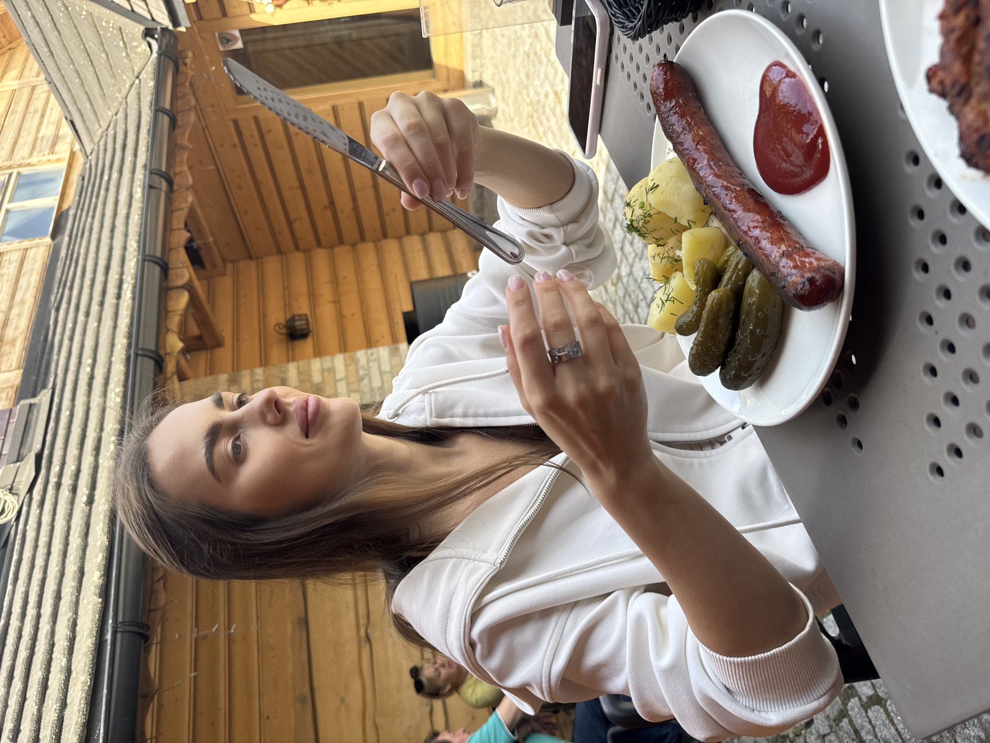 A woman is seated at an outdoor table, holding a knife above a plate of food. The plate contains a sausage, potatoes with dill, pickles, and a dollop of ketchup. She is wearing a white hooded jacket and has rings on her fingers. The table is a dark, perforated metal. Behind her are wooden walls and roof of a building. The location is outdoors in an unspecified urban environment.