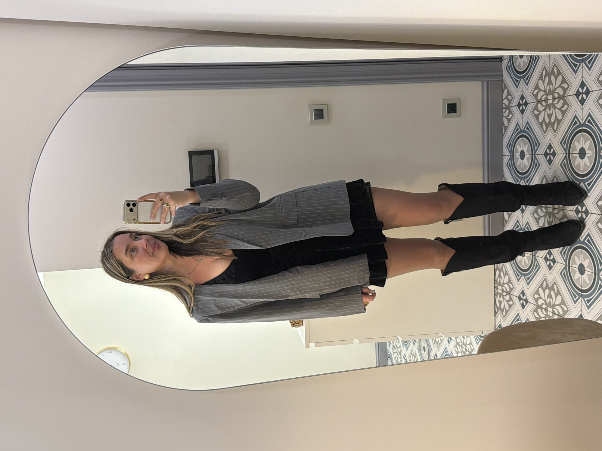 A woman is taking a selfie in a mirror. She is wearing a black dress with a gray pinstripe blazer and black boots. The reflection shows a room with a clock on the wall, a television, light switches, and tiled flooring. It is not possible to determine the location to be in Moscow, Russia.