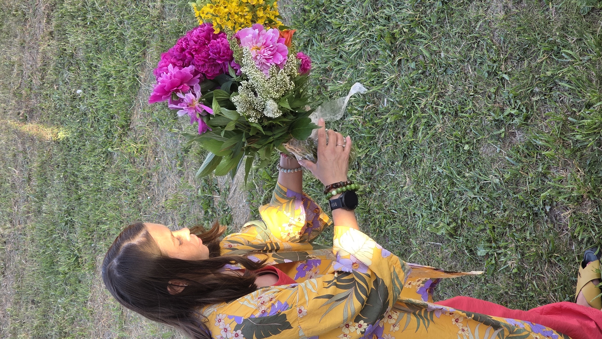 A woman with long brown hair, wearing a yellow kimono with a floral pattern and a red undergarment, is kneeling in a grassy area. She is holding a bouquet of flowers, which includes pink peonies, white flowers, and yellow flowers. Her hands are wrapped around the stems of the bouquet, and she appears to be arranging them. She is wearing bracelets on both wrists, one with green beads and the other with a mix of green and brown beads, and a black smartwatch on her left wrist. A ring is visible on her right hand. The background consists of green grass. The scene is illuminated by natural sunlight, casting a shadow to the right of the woman.